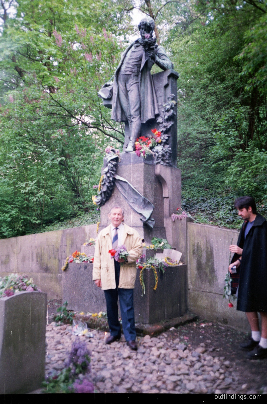 Monumental bronze statue of a robed figure in a park setting, surrounded by floral tributes. A man in a beige coat holds a bouquet of red roses, while another person in dark attire stands nearby. Gravestone details and lush greenery frame the scene, suggesting a solemn tribute. Likely Eastern European, 1980s–1990s era.