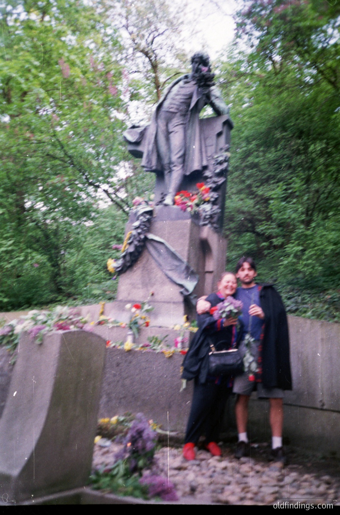 Two individuals pose beside a solemn bronze statue of a seated figure draped in robes, likely a historical or cultural monument. Surrounding the statue are floral tributes—wreaths and bouquets—suggesting a memorial or tribute site. Lush greenery frames the scene, indicating an outdoor park or cemetery setting. The attire and photo quality suggest a mid-20th-century timeframe. [Two people standing beside a solemn statue adorned with floral tributes in a lush outdoor setting, likely a memorial site from the 1960s–1980s]