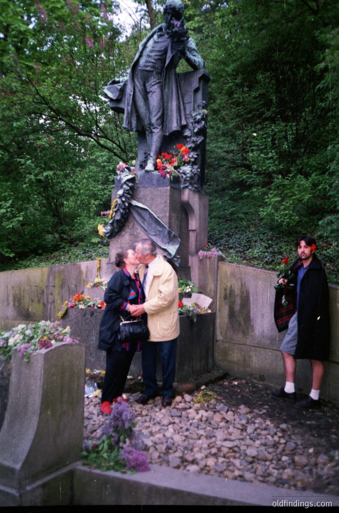 Two individuals place flowers at a solemn monument in a wooded area, surrounded by grave markers and greenery. The statue depicts a robed figure in contemplative pose, likely a historical or cultural figure. Mid-20th century attire suggests a or setting. Potential European cemetery or memorial site.