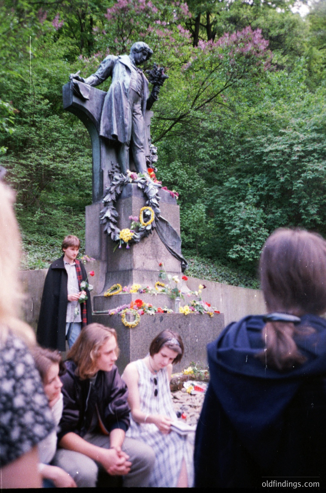 Bronze statue of seated figure in classical attire, surrounded by floral wreaths and bouquets at a memorial site. Group of people in 1980s/90s attire paying respects. Lush greenery and blooming trees in background. Likely Eastern European public square.