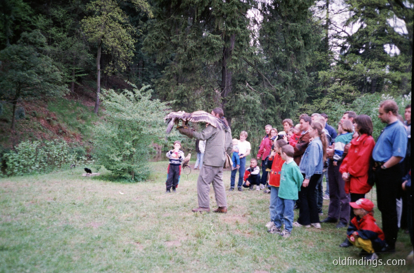 A group of people in a forested area, likely a nature reserve or park, observing a bird-feeding demonstration. The central figure feeds a bird from a bag or pouch, while onlookers—adults and children—watch intently. Casual outdoor attire suggests a family-friendly event, possibly educational or recreational. Dense evergreen trees frame the scene, indicating a temperate climate. Image likely from late 20th century (1980s–1990s) based on clothing styles.