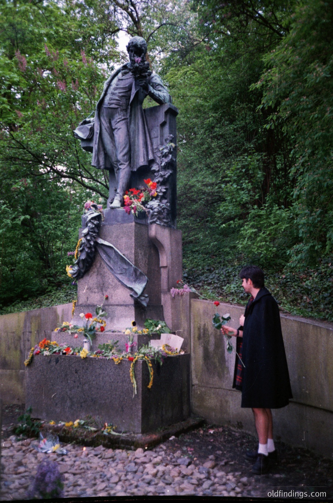 Neoclassical statue of a robed figure in contemplative pose, adorned with floral wreaths and red roses at its base. A person in black attire places a single red rose on the pedestal. Lush greenery surrounds the monument, suggesting an outdoor park setting. Likely Eastern European, 20th-century memorial style.