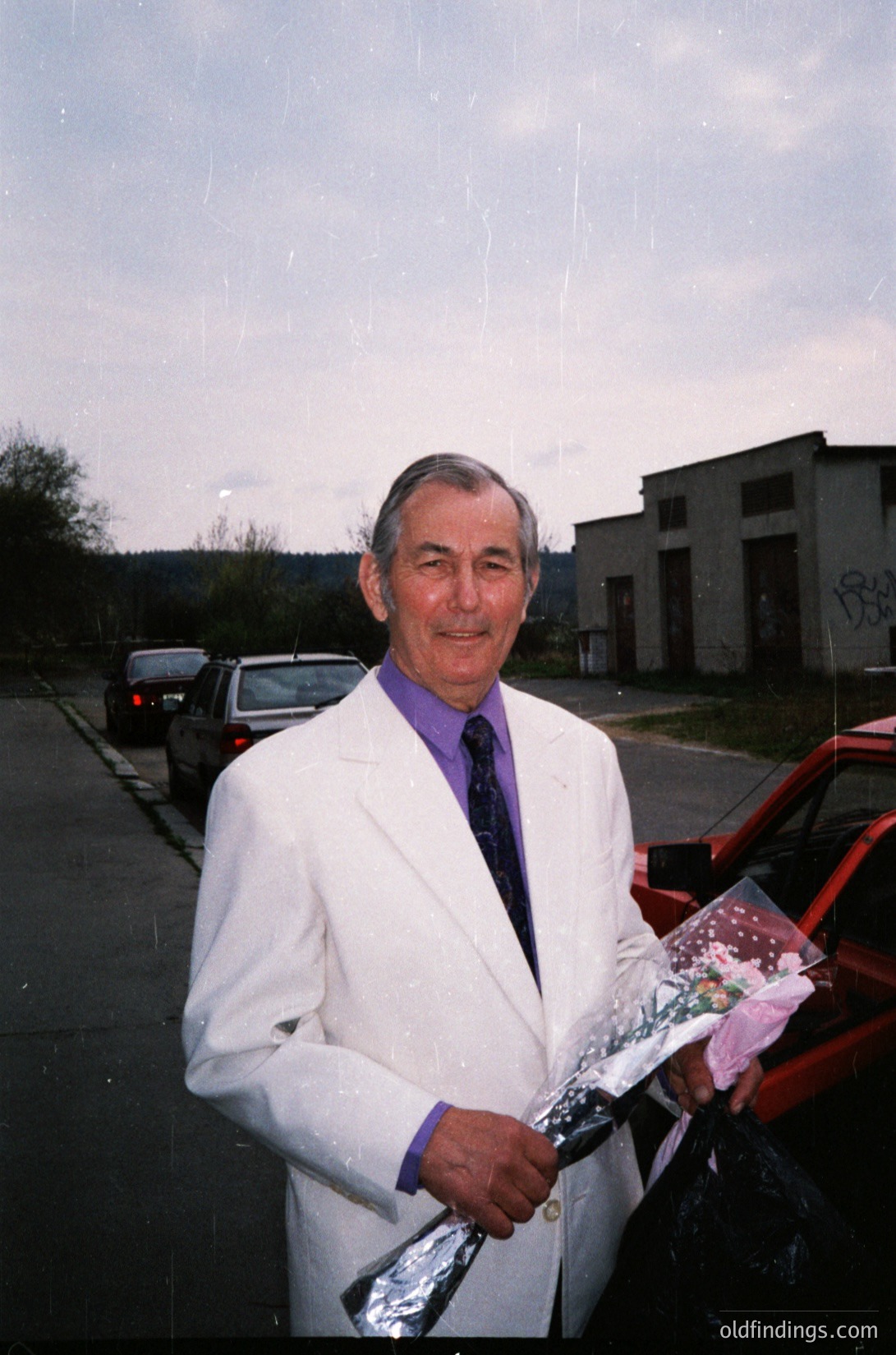 Mid-20th century man in formal attire (white suit, purple tie) holding a wrapped gift, likely a celebratory occasion. Urban industrial background with parked cars and graffiti-marked buildings.