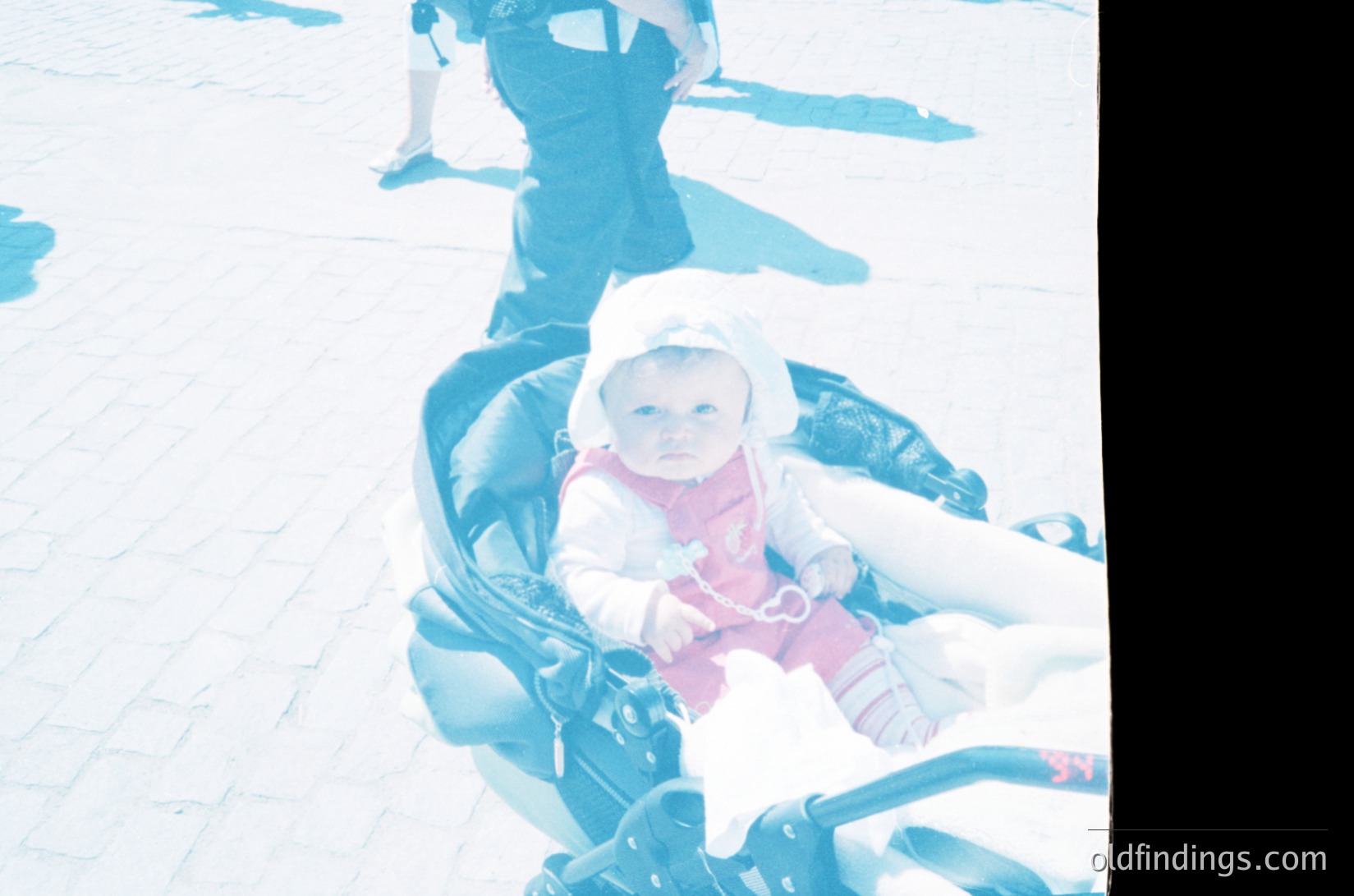 Vintage sepia-toned photo of an infant in a classic stroller on a paved surface, likely mid-20th century. Child wears a white hat and pink jacket, seated in a black metal stroller with a padded seat. Background shows blurred figures, suggesting an outdoor urban or park setting.