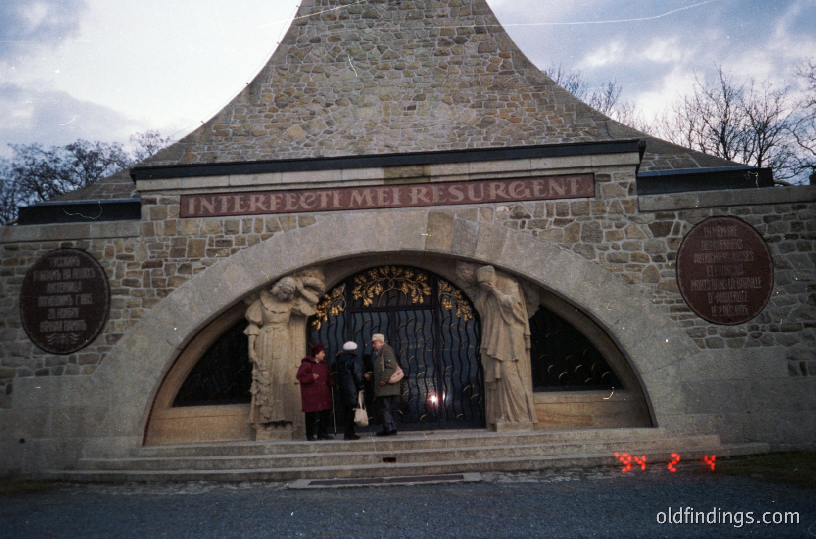 Stone archway entrance with Latin inscription *"Interfecti Mel Resurgent"* (Martyrs Rise Again) flanked by sculpted figures. Two elderly individuals in winter coats pose under the arch, with a plaque to the right. Rustic stonework and vintage color tone suggest mid-20th century. Likely a memorial or cemetery gate.