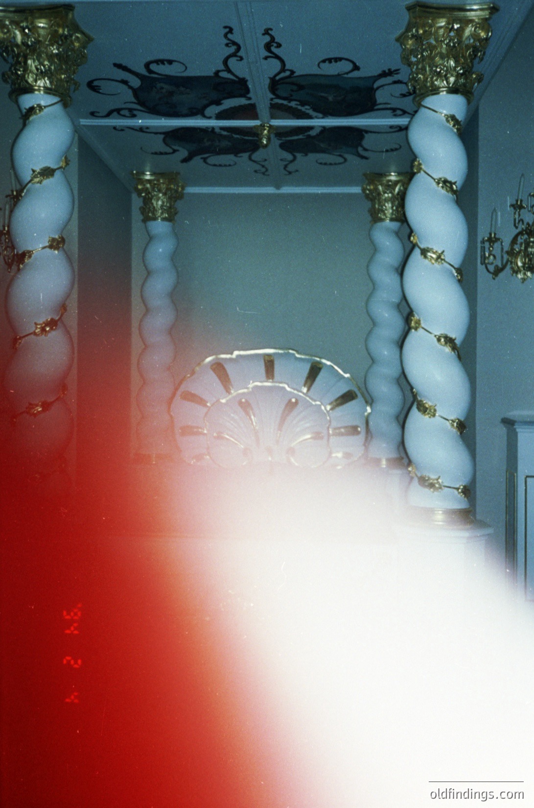 Vintage interior shot featuring ornate blue-and-gold striped columns framing a grand staircase. Circular skylight above reveals architectural details. Warm lighting casts dramatic shadows. Likely 1960s–1980s Eastern European hotel or public building.