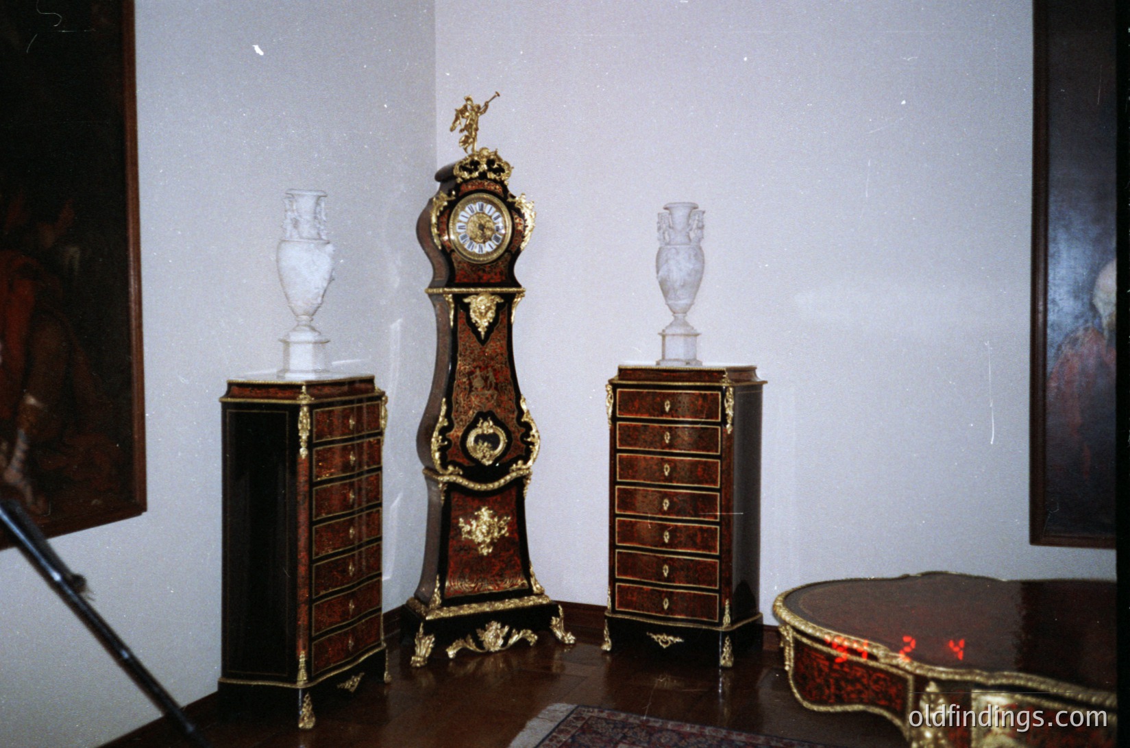 Antique Rococo-style furniture ensemble featuring ornate wooden cabinets with gold accents, paired with two white marble urns and a grand clock. Intricate carvings and gilded details highlight 18th-century craftsmanship. Likely from a European aristocratic interior, possibly France or Germany.