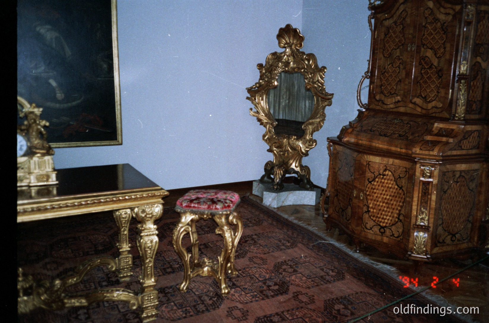 Baroque-style interior featuring ornate gold-accented furniture: intricately carved wooden cabinet with floral motifs, gilded mirror frame on a marble base, and a small red velvet stool on clawed legs. Rich red patterned carpet contrasts with muted walls. Likely European palace or aristocratic residence, 17th–18th century.