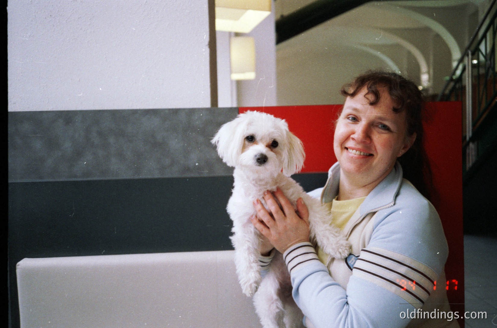 Vintage candid of a woman holding a small white poodle in an indoor setting, likely a public transport hub. She wears a striped sweater with a fur-lined collar, and the station’s curved seating and red/black/white striped wall suggest mid-20th-century design. Warm lighting and candid expression evoke nostalgia.