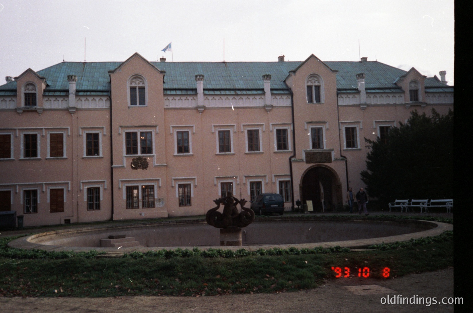 Neoclassical building with symmetrical façade, featuring arched windows and a central entrance. Prominent statue in circular courtyard with red-tinted glass overlay (date stamp: 1993.10.8). Overcast sky, minimal greenery.