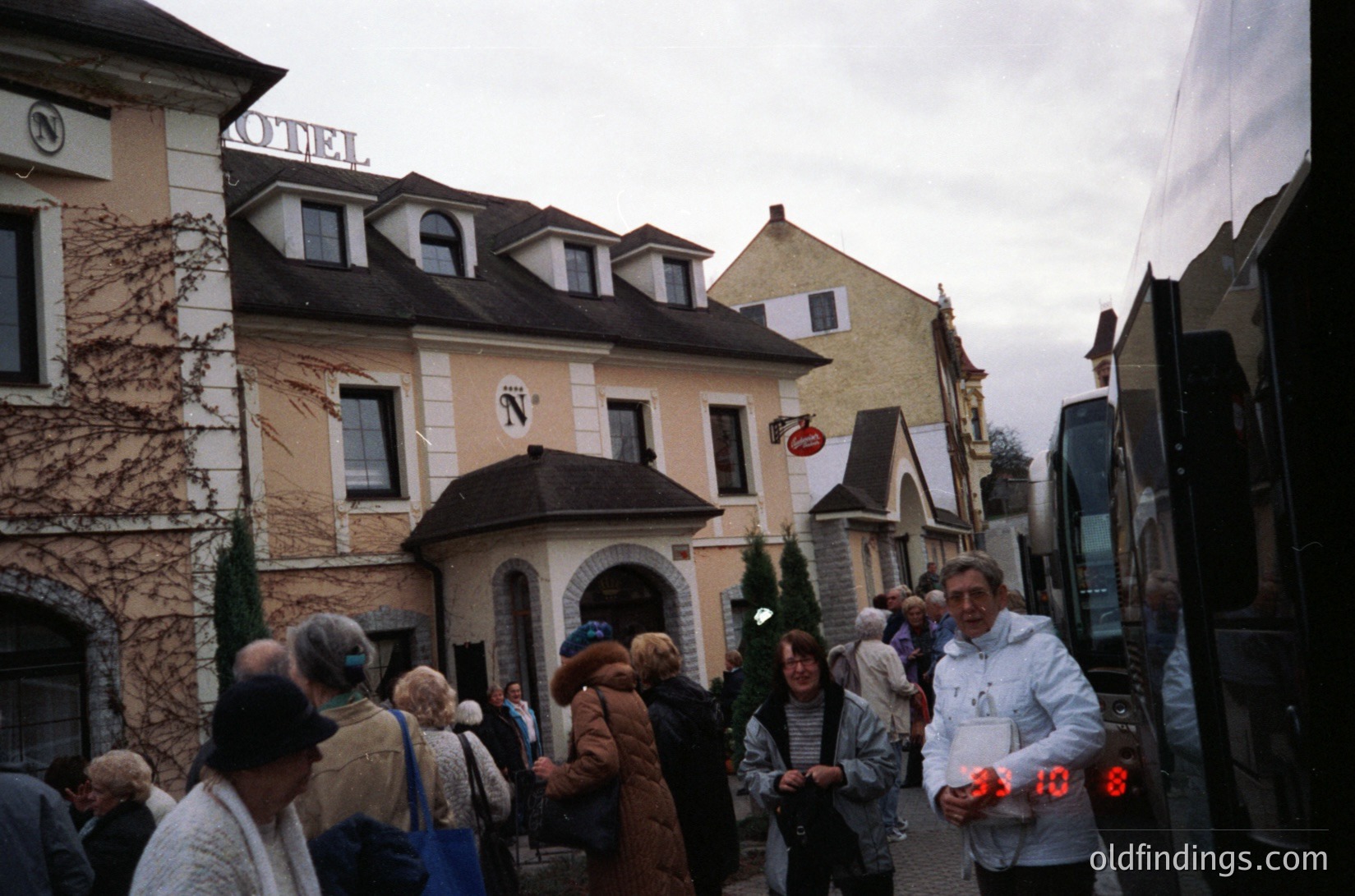 Vintage European street scene featuring a bustling crowd near a **1970s-era hotel** with "N" signage. Ivy-covered façade, gabled roofs, and a clock displaying **9:10**. Tourists in winter coats, some carrying luggage, gather outside. Overcast sky and parked buses suggest a transit hub.