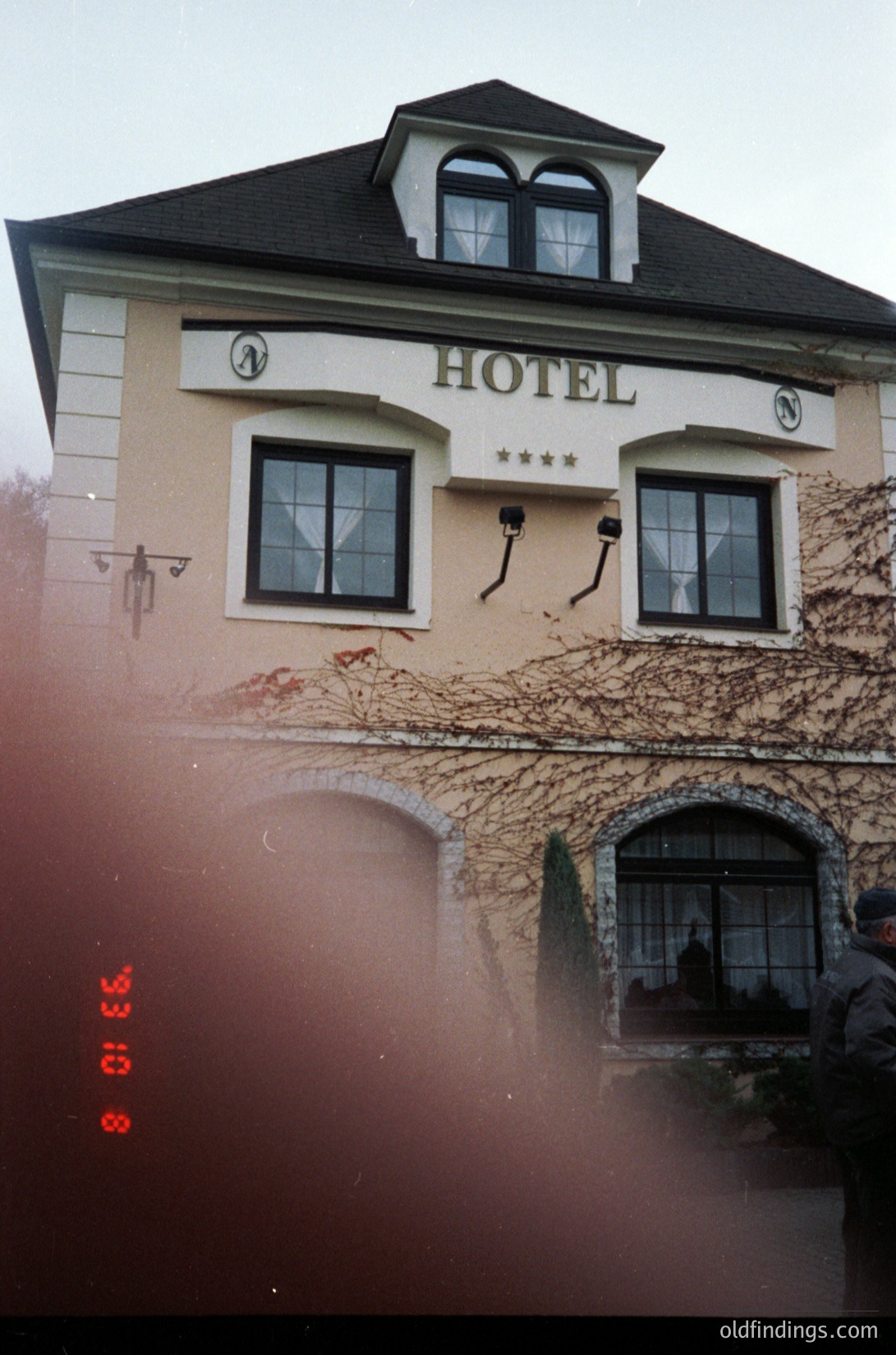 Vintage 35mm photo of a European-style hotel façade with ivy-covered walls, arched windows, and a peaked roof. Decorative clock faces flank the sign, and a balcony with a wrought-iron railing sits below. Film grain and red date stamp (1993) visible.
