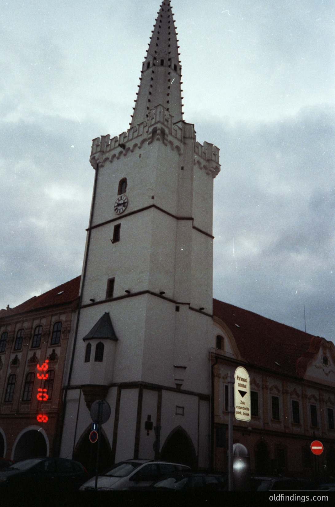 Historic Gothic-style tower with pointed spire, likely a church or town hall, featuring crenellated upper sections and a coat-of-arms emblem. Surrounding buildings exhibit Baroque architectural details, including arched windows and decorative cornices. Urban street scene with parked cars and street signs ( )