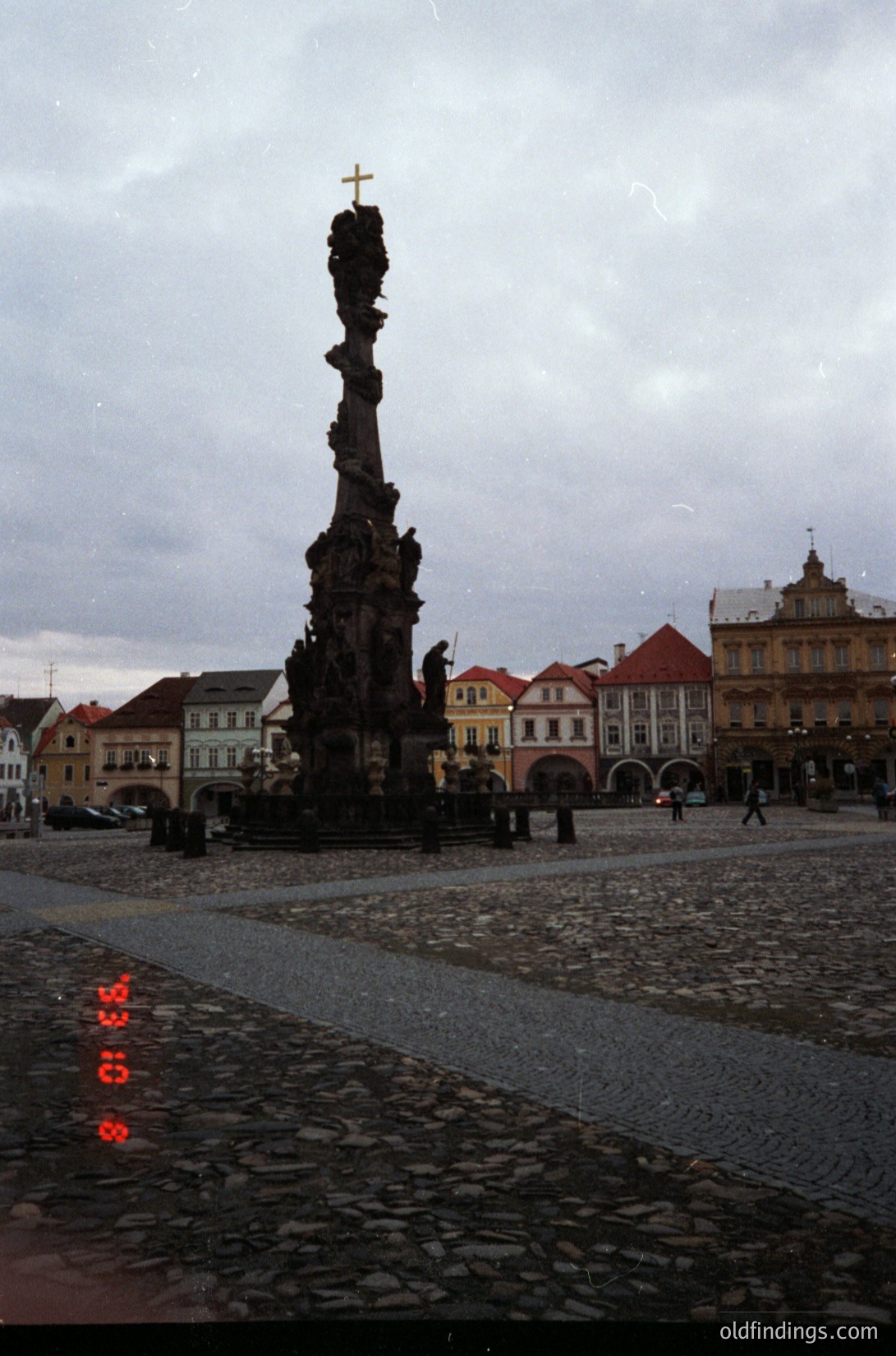Baroque-style column monument with crucifixion scene atop, set on cobblestone square. Surrounding historic European architecture with arched windows and gabled roofs. Likely Czech Republic, 18th–19th century.