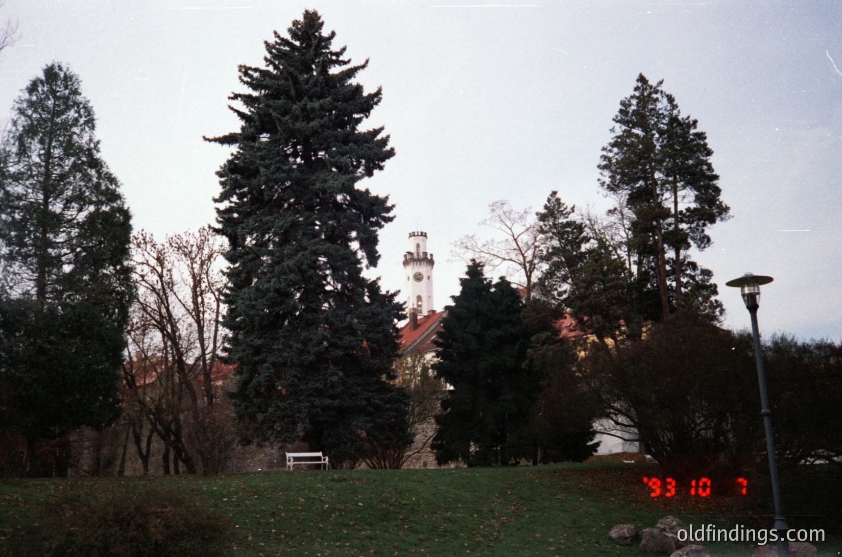 Vintage photo of a park-like setting with a historic tower in the background, likely European. The date stamp "93 10 7" suggests October 1993. Tall evergreen trees frame the scene, and a vintage-style lamp post with a red "7" is visible. The tower features a clock and a red roof.