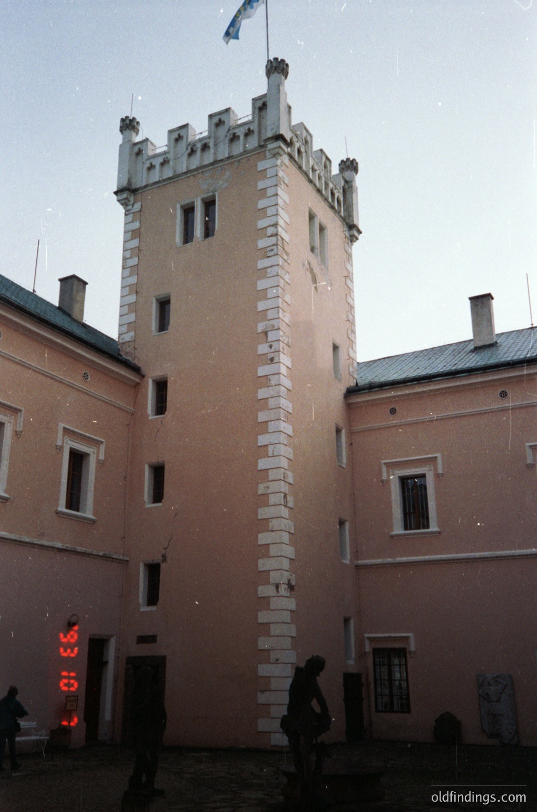 Historic courtyard building with Gothic Revival tower featuring crenellated parapets and vertical stone accents. Light-colored facade contrasts with muted pink walls. Overcast sky and minimal winter activity suggest cold weather. Likely Eastern European institutional or cultural site.