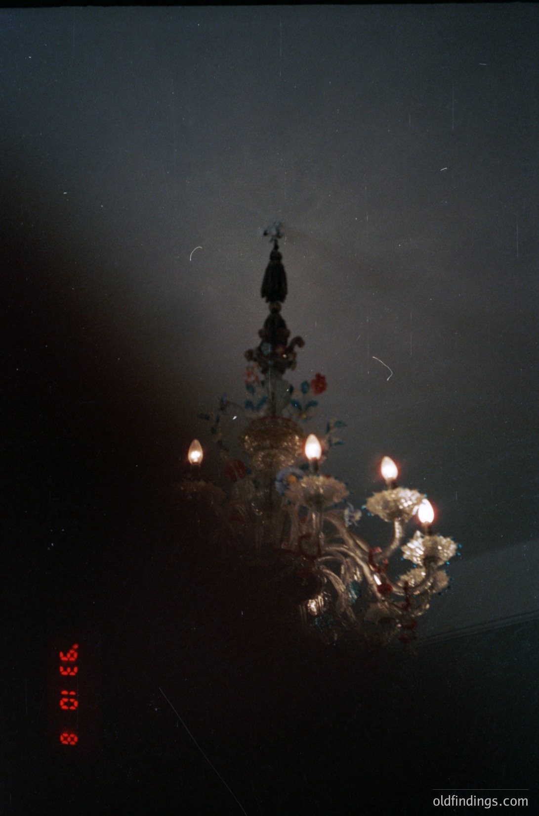 Vintage close-up of ornate chandelier with intricate metalwork, floral motifs, and warm bulbs. Dark, moody lighting enhances details. Likely 1990s indoor setting, possibly a restaurant or hotel lobby.