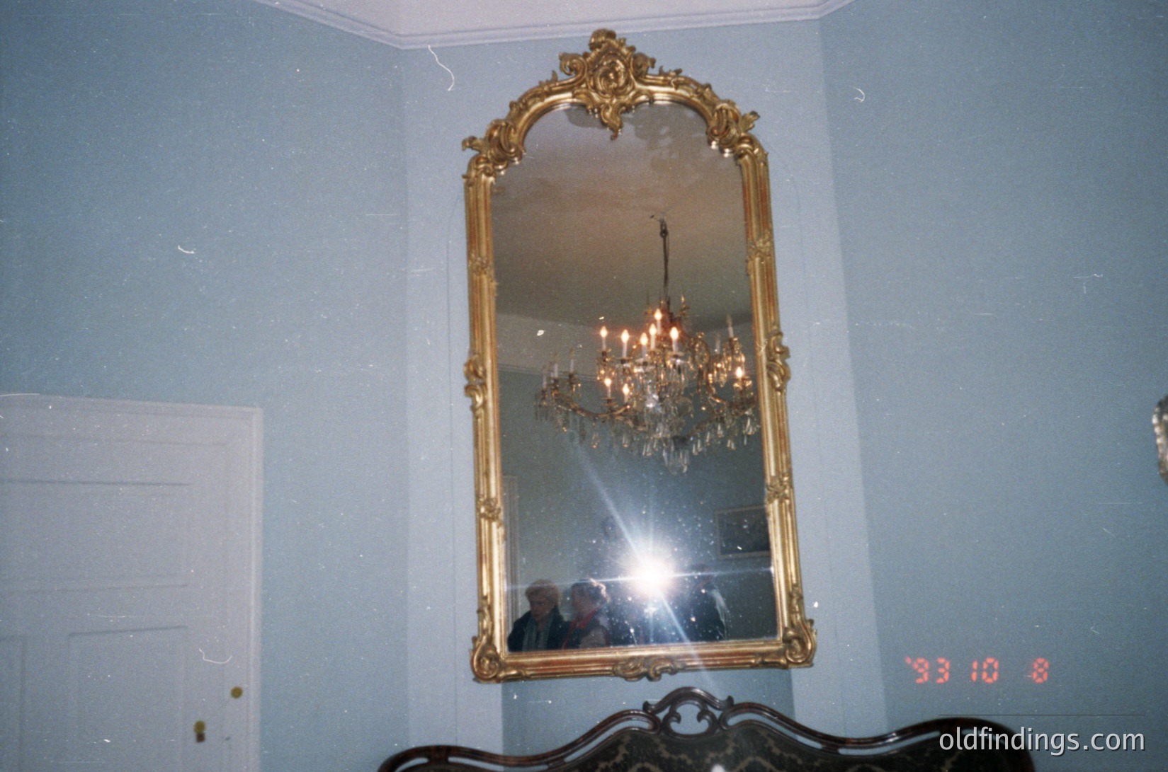 Vintage ornate gold-framed mirror reflecting a chandelier in a dimly lit room. Classic interior design with intricate floral motifs. Likely 1970s–1980s European or American residential setting. Ideal for vintage decor, interior design references, and historical home studies.