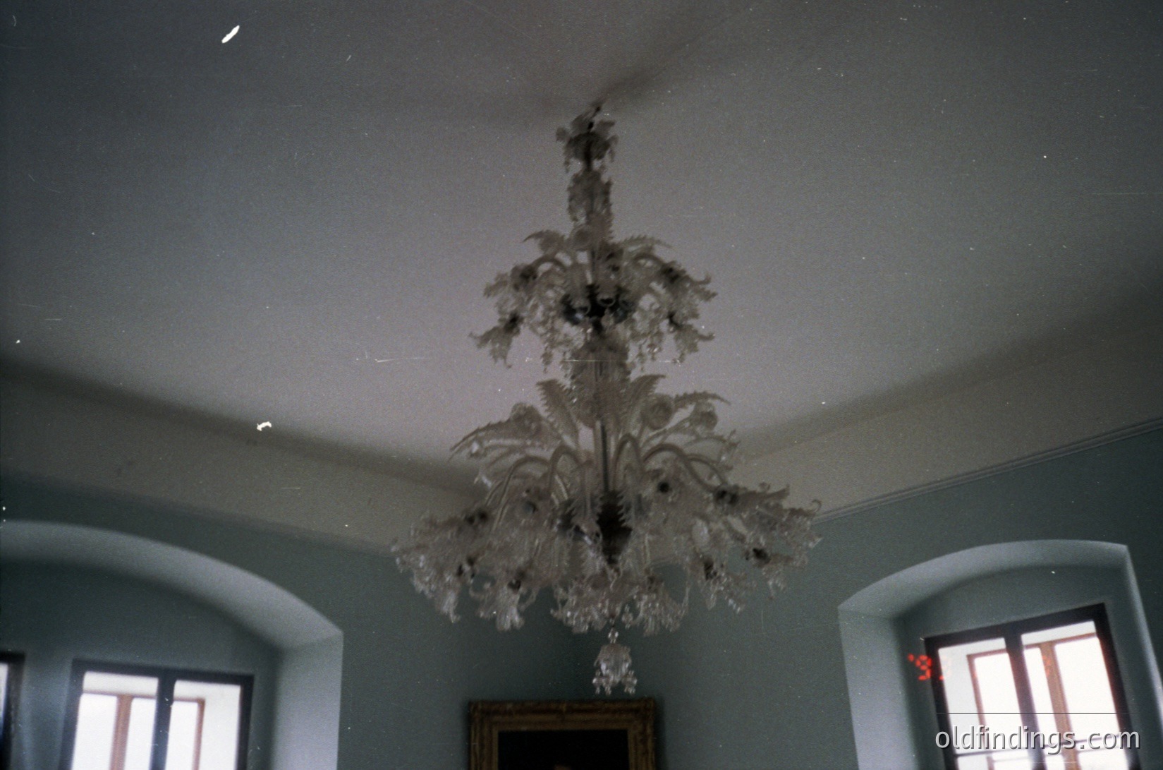 Vintage chandelier with ornate crystal branches suspended in a dimly lit, classical-style room. Symmetrical arched windows with wooden frames flank the space, allowing natural light. Likely mid-20th century European interior, possibly .
