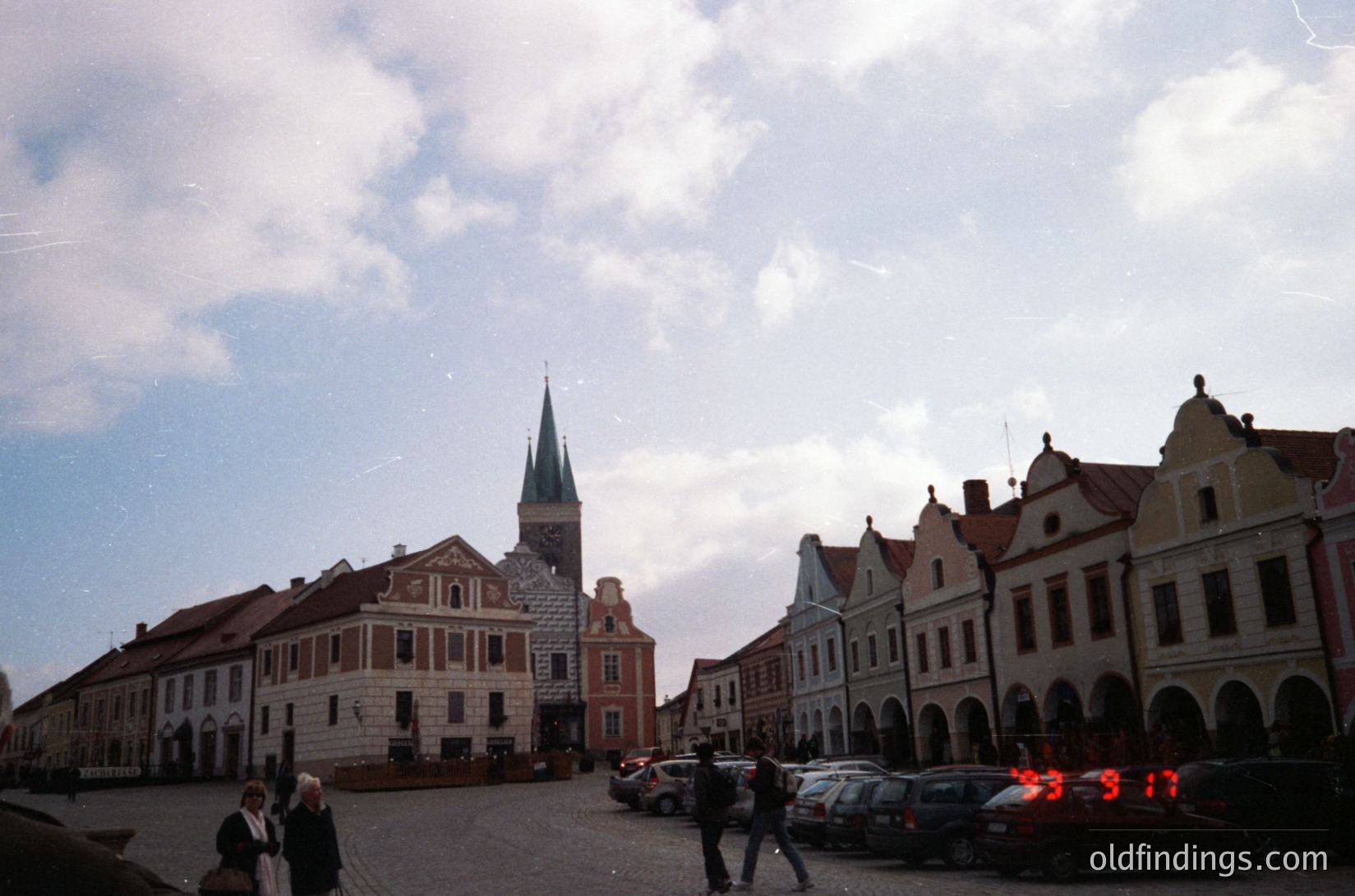 Historic European town square with Renaissance-style buildings featuring stepped gables and arched ground floors. Central church tower with a green spire dominates skyline. Cobblestone plaza with parked cars and pedestrians. Likely or , -2000s.