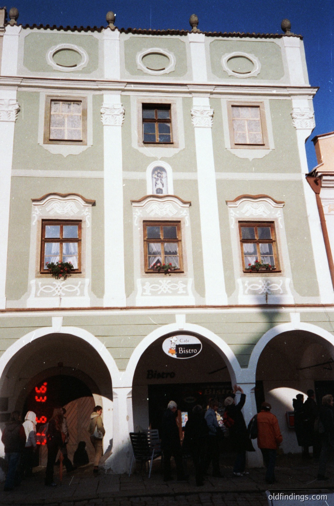 Three-arched entrance of a **Baroque-style building** with cream stucco and decorative moldings. Ground-floor signage reads "Bistro" with a logo. Crowd of casually dressed people entering/exiting. Symmetrical windows with flower boxes. Likely **Central/Eastern European** (1990s–2000s).