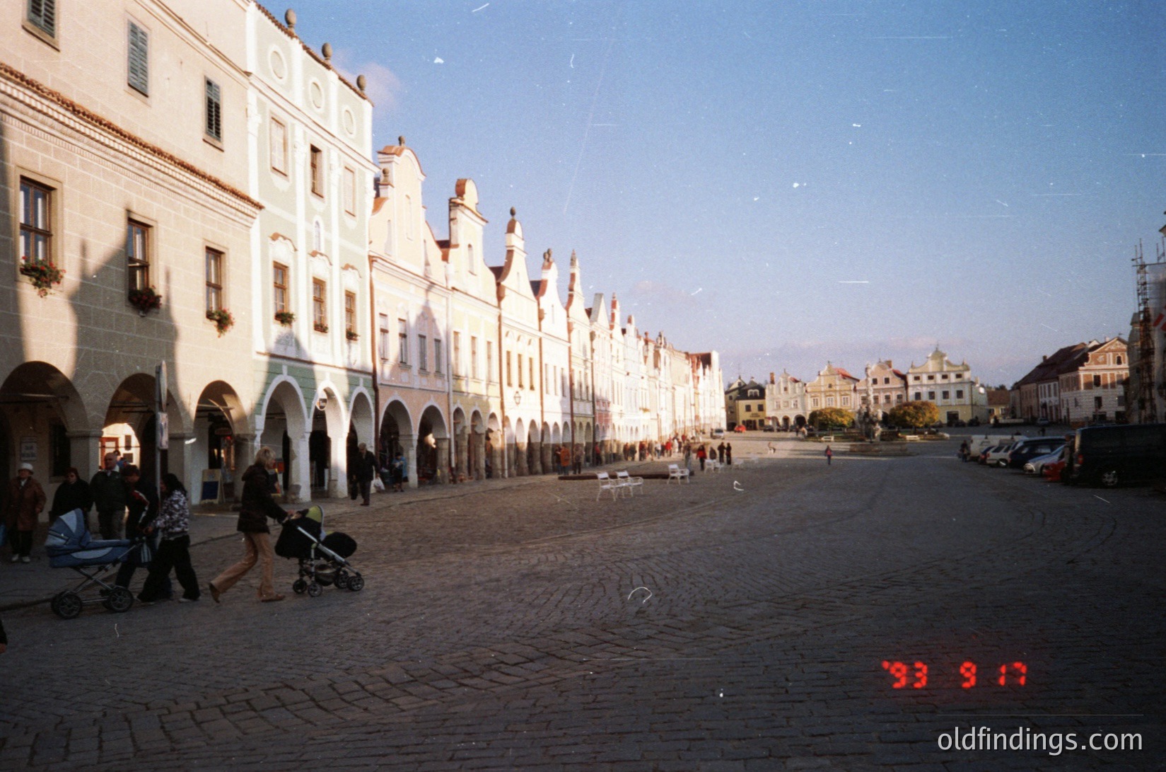 Historic European plaza with Renaissance-style arcaded buildings, likely 1993. Cobblestone street lined with whitewashed facades featuring pointed gables and arched walkways. People strolling with strollers, parked cars, and vintage date stamp (9/17).