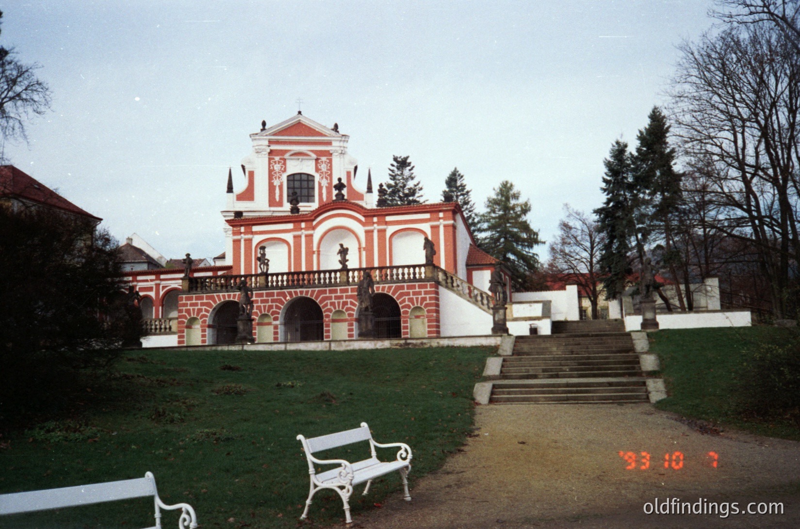 Baroque-style pavilion with red-and-white striped brickwork, central dome, and arched colonnades. Overgrown park setting with bare trees and a white bench in foreground. Likely Eastern European, mid-20th century.