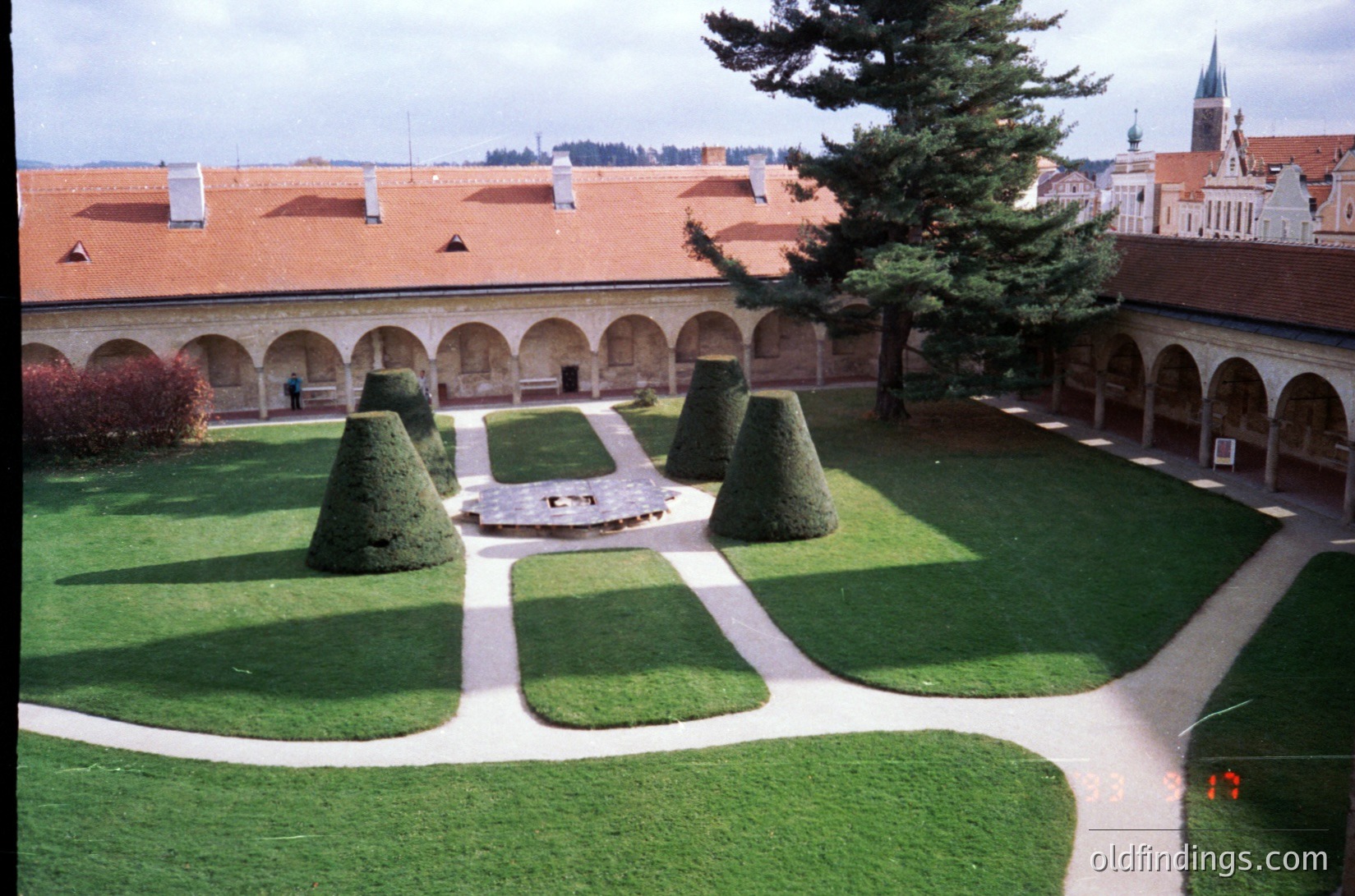 Neoclassical cloister courtyard with symmetrical topiary gardens, gravel pathways, and arched colonnades. Red-tiled roofs and historic church spires visible in background. Likely European, possibly , with formal or influences. --- *Note: Exact location indeterminate but stylistic cues suggest 18th–19th century European monastic or institutional architecture.*