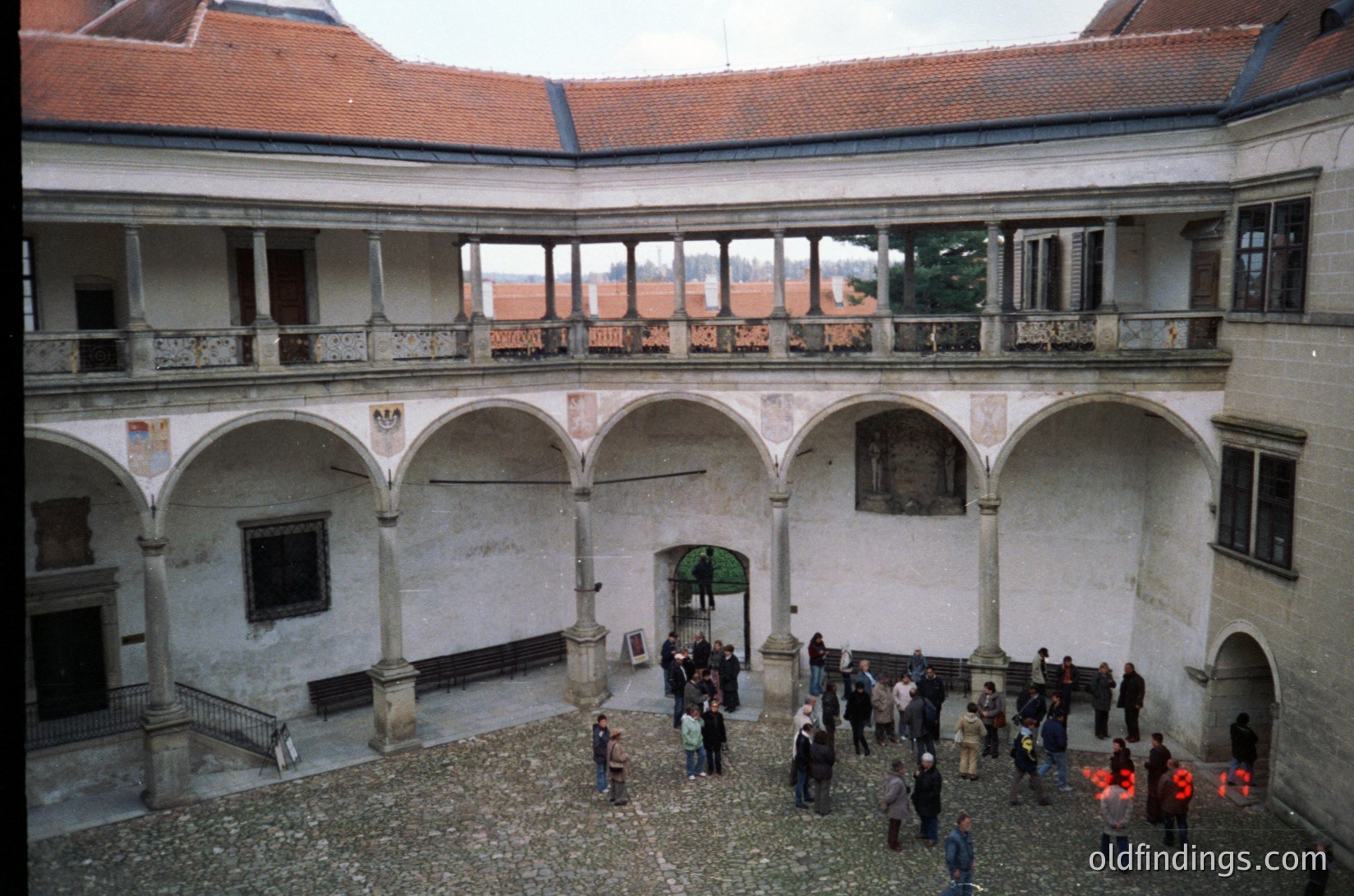 Two-story courtyard with arched colonnades, red-tiled roof, and weathered plaster walls. Group of people gathered near central archway. Likely European historic building, possibly 19th–20th century.