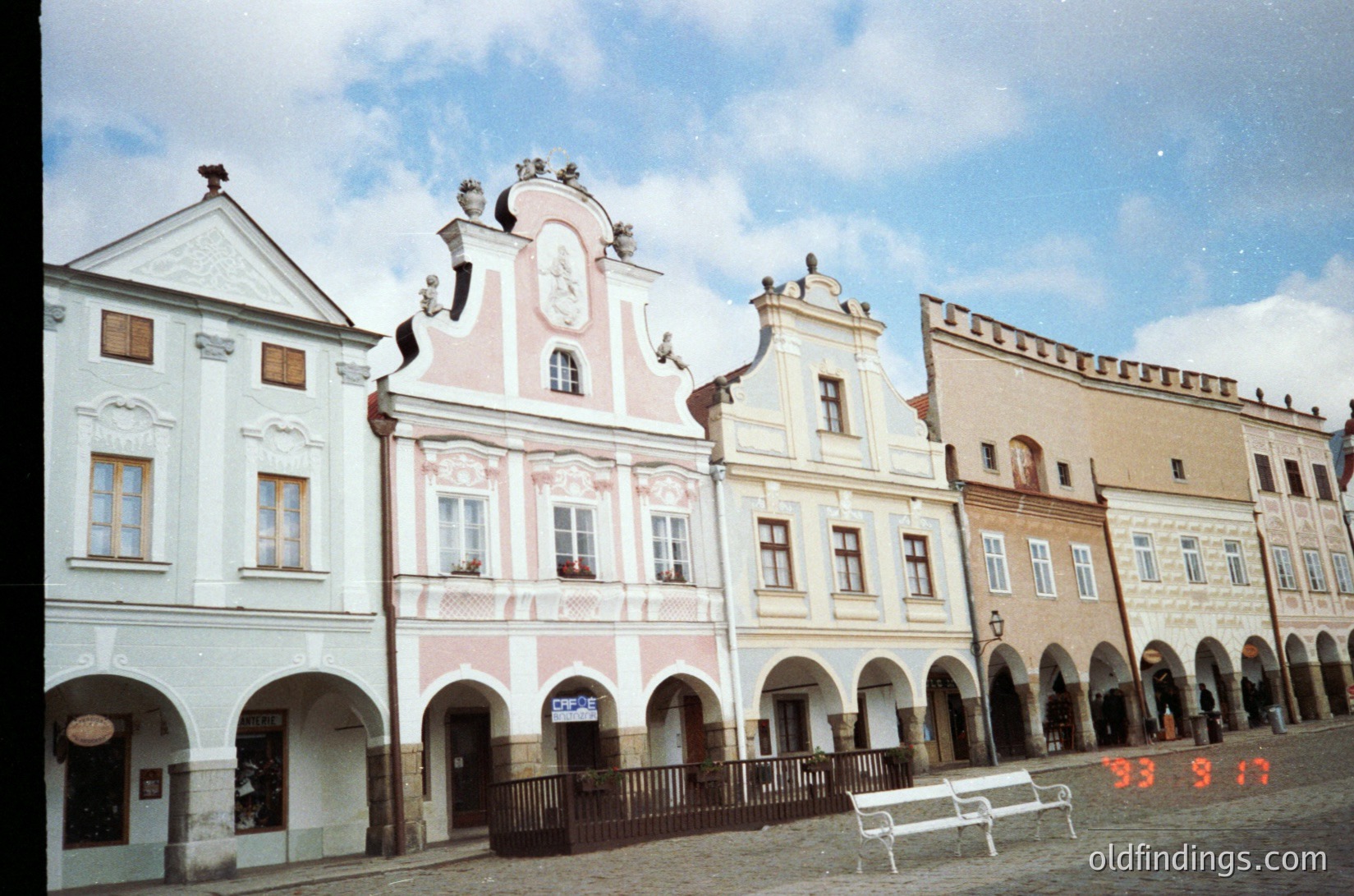 Baroque-style town square featuring three-story buildings with arched ground floors, decorative facades, and ornate rooftops. Central building showcases intricate stucco reliefs and a clock. Cobblestone plaza with a bench and pedestrians. Likely European, 18th–19th century.