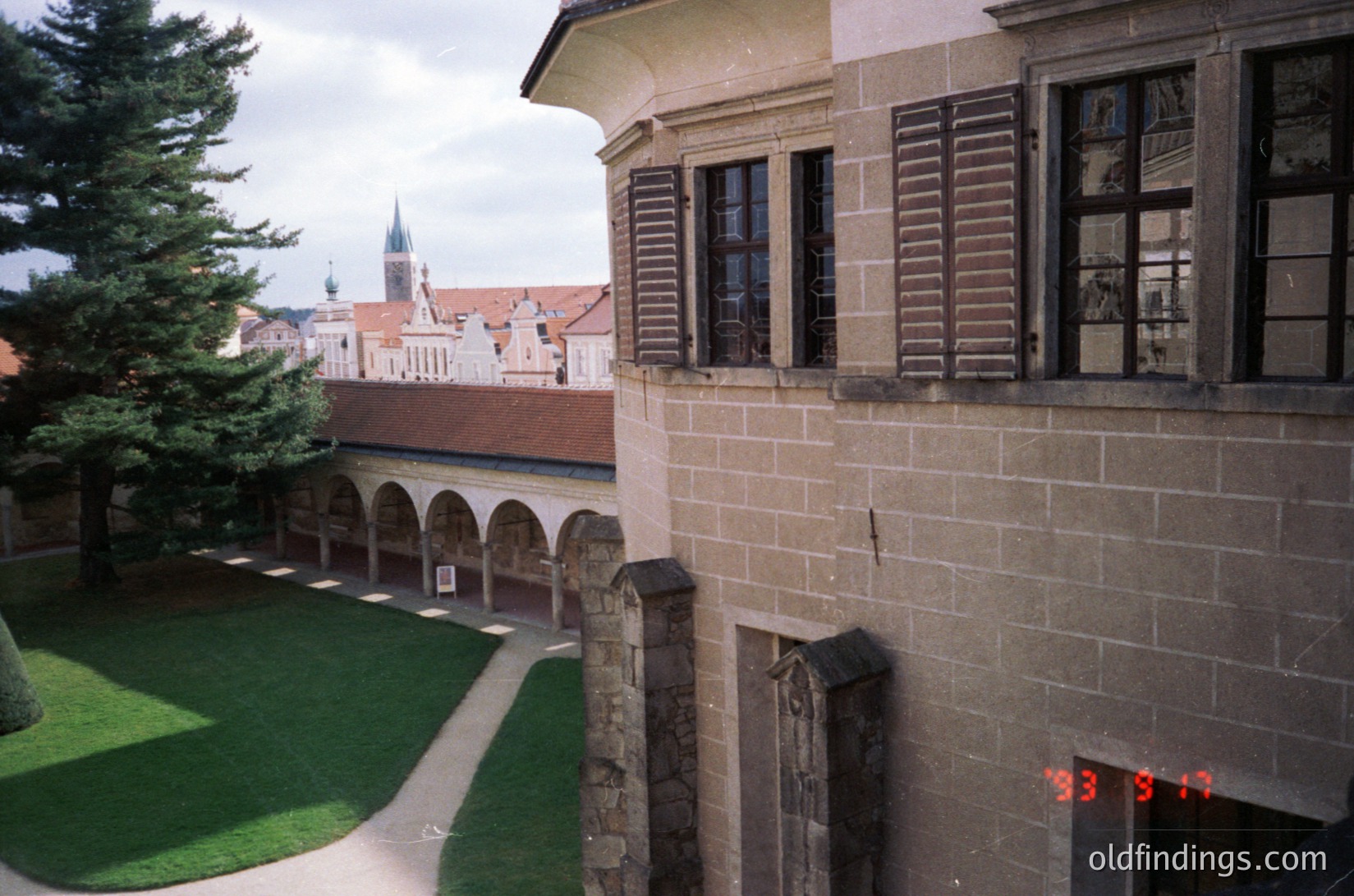 Historic courtyard view of a stone building with arched colonnades, likely a monastery or cloister. Gothic-style church spire visible in background. Overgrown grass and dated stamp (1993) suggest mid-1990s Eastern Europe. Architectural details include shuttered windows and carved stonework.