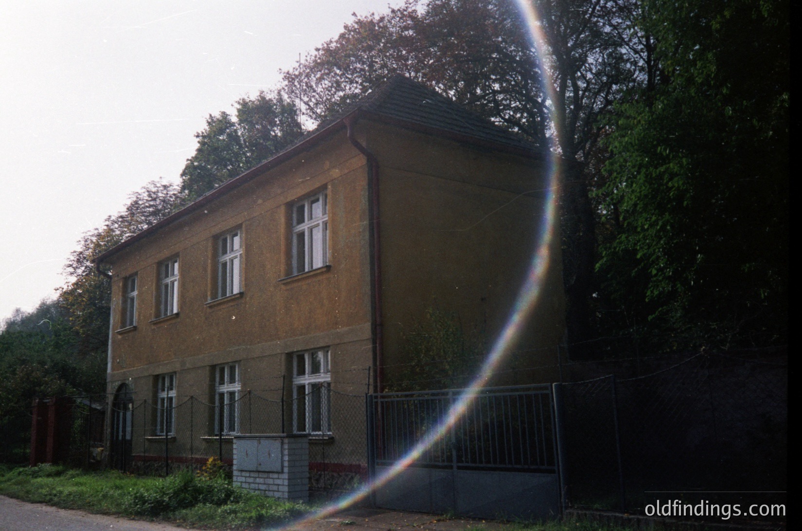 Two-story brick building with symmetrical windows, surrounded by chain-link fence. Lens flare obscures part of the frame. Likely Eastern European architecture, mid-20th century.