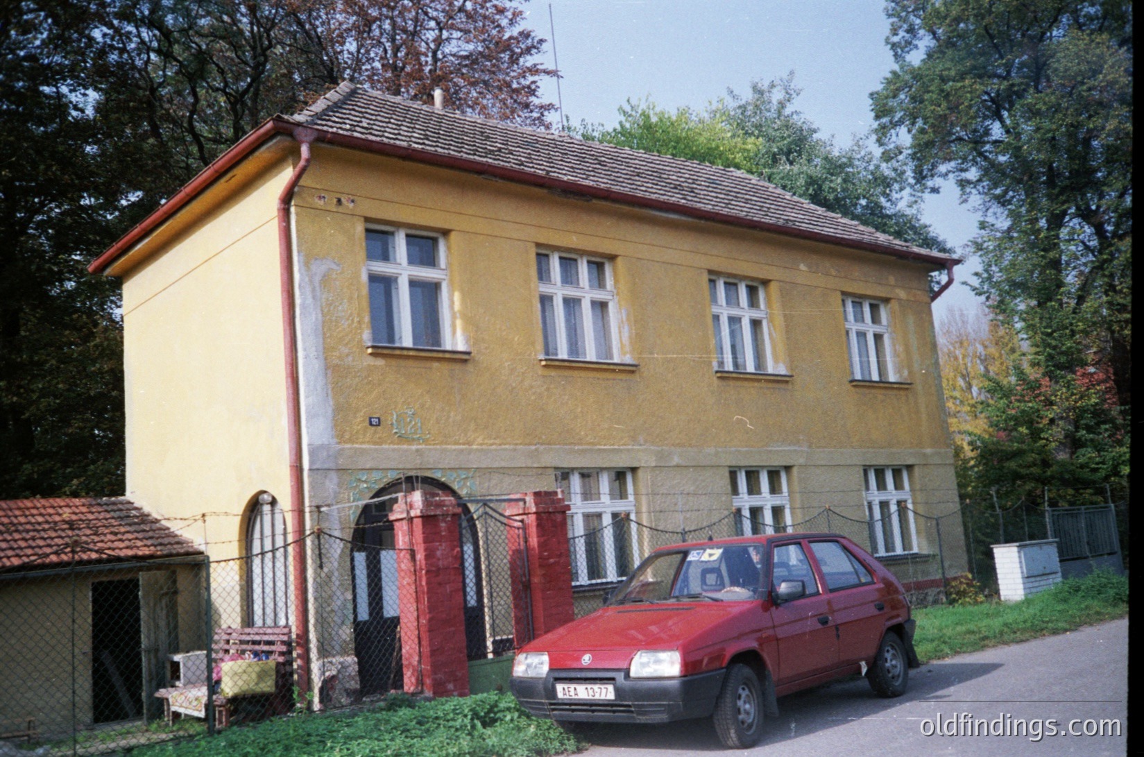 Two-story yellow residential building with red accents, likely from the 1970s-1980s. Peeling exterior paint and faded red door frame indicate aging. Red hatchback car parked in front suggests mid-late 20th-century Eastern Bloc region. Surrounding greenery and chain-link fence hint at suburban or semi-rural setting.
