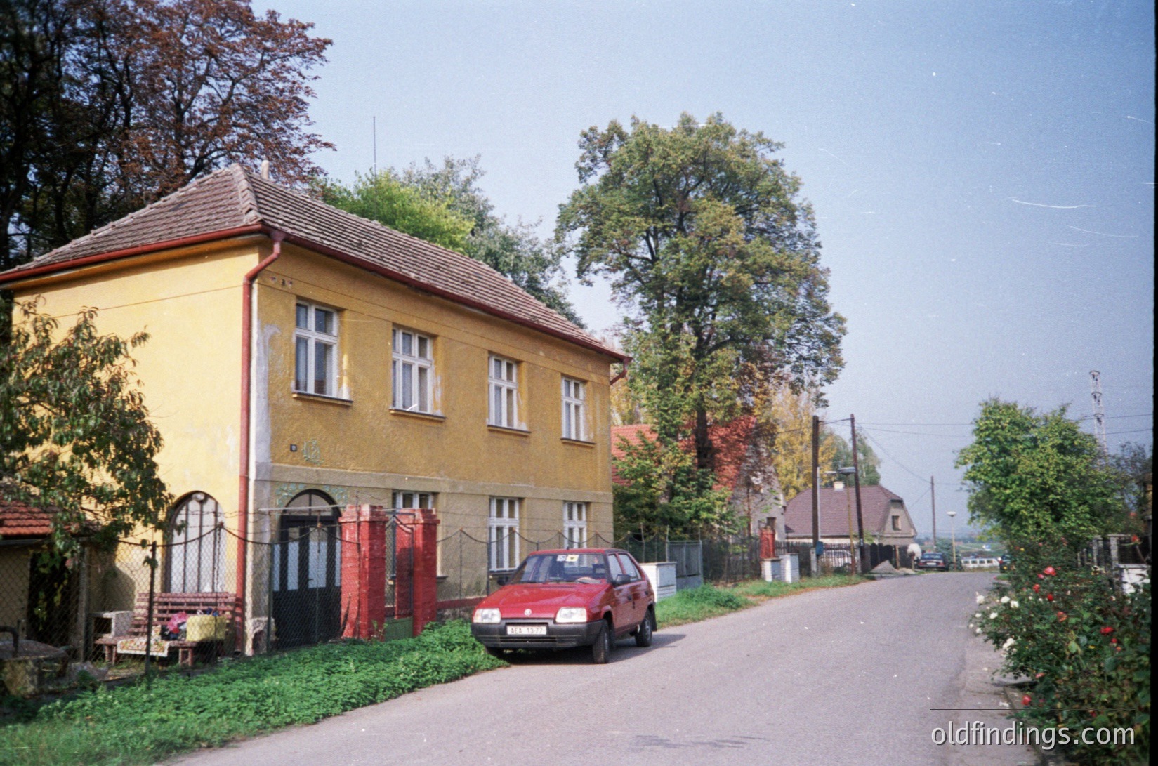 Vintage 1980s-era residential street with Soviet-era architecture: two-story yellow building featuring red garage doors and white-framed windows. A red hatchback car parked on a narrow, tree-lined road. Overgrown greenery and autumn foliage on both sides. Rural or suburban setting, likely Eastern Europe.