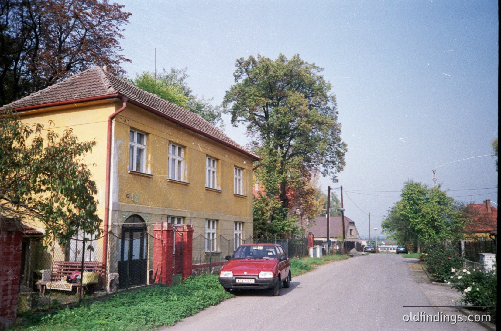Two-story yellow residential building with red-tinted window frames and a peaked roof, flanked by red curtains. A red car parked beside a chain-link fence. Quiet suburban street with mature trees and overgrown greenery. Likely Eastern European architecture, mid-20th century.