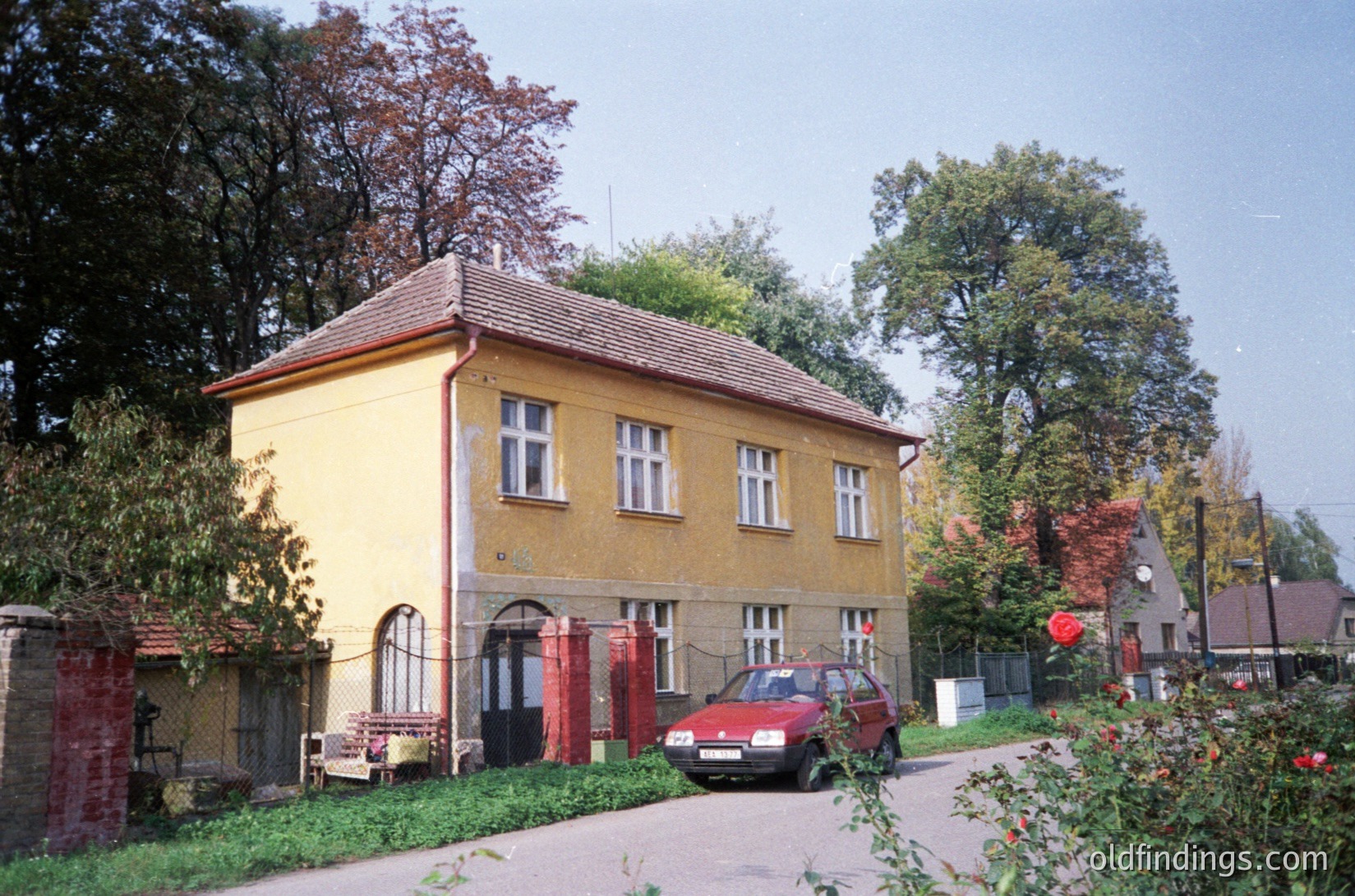Two-story yellow residential building with red trim, featuring arched entryway and white-framed windows. Red car parked on paved driveway beside chain-link fence. Autumn foliage in foreground and background. Likely Eastern European suburban setting, mid-20th century architecture.