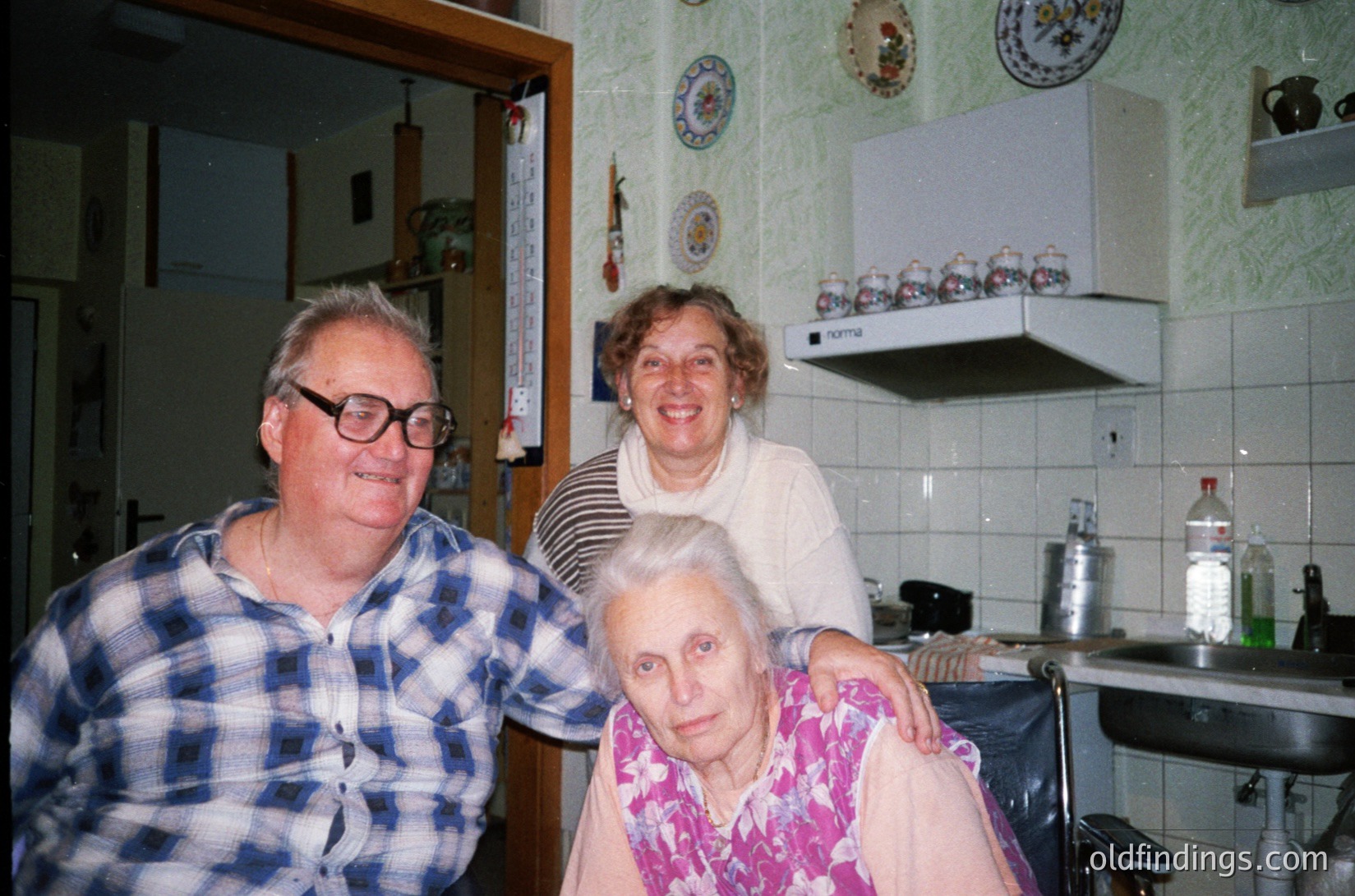 Mid-20th century kitchen interior with three elderly individuals posing. Man in checkered shirt, woman in striped sweater, seated woman in floral blouse. White-tiled walls adorned with decorative plates and a white range hood. Likely *(Note: "Bulgaria" is inferred from cultural elements; adjust if location differs.)*