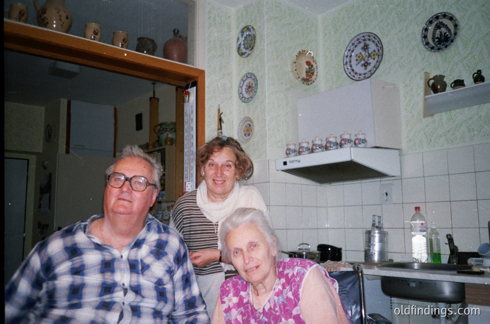 Three elderly individuals pose in a vintage kitchen, likely Eastern Europe, 1980s–1990s. White-tiled walls feature decorative plates and ceramic figurines. The man wears a plaid shirt; the seated woman sports a floral blouse. Functional appliances and shelves suggest a lived-in, family-centric space.