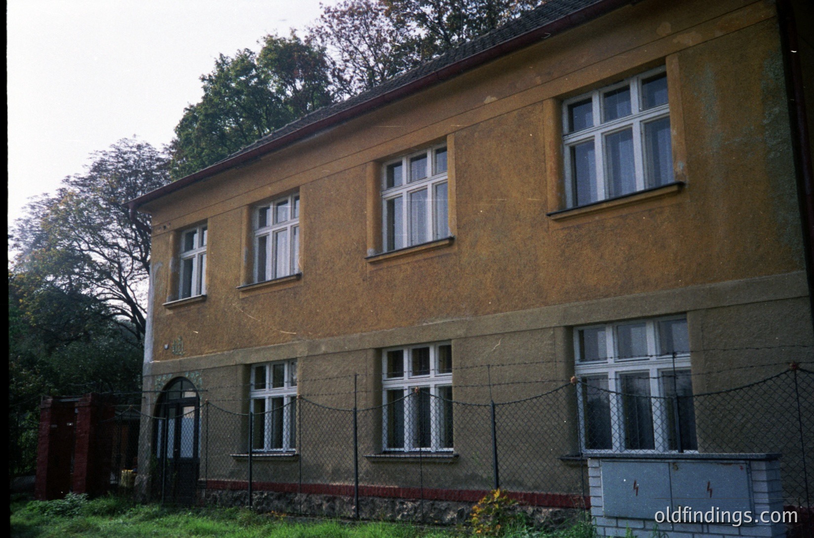 Two-story brick building with aged yellowish facade, featuring symmetrical white-framed windows. Chain-link fence with red brick base and metal utility box at ground level. Overgrown greenery and trees in background. Likely Eastern European residential architecture, mid-20th century.