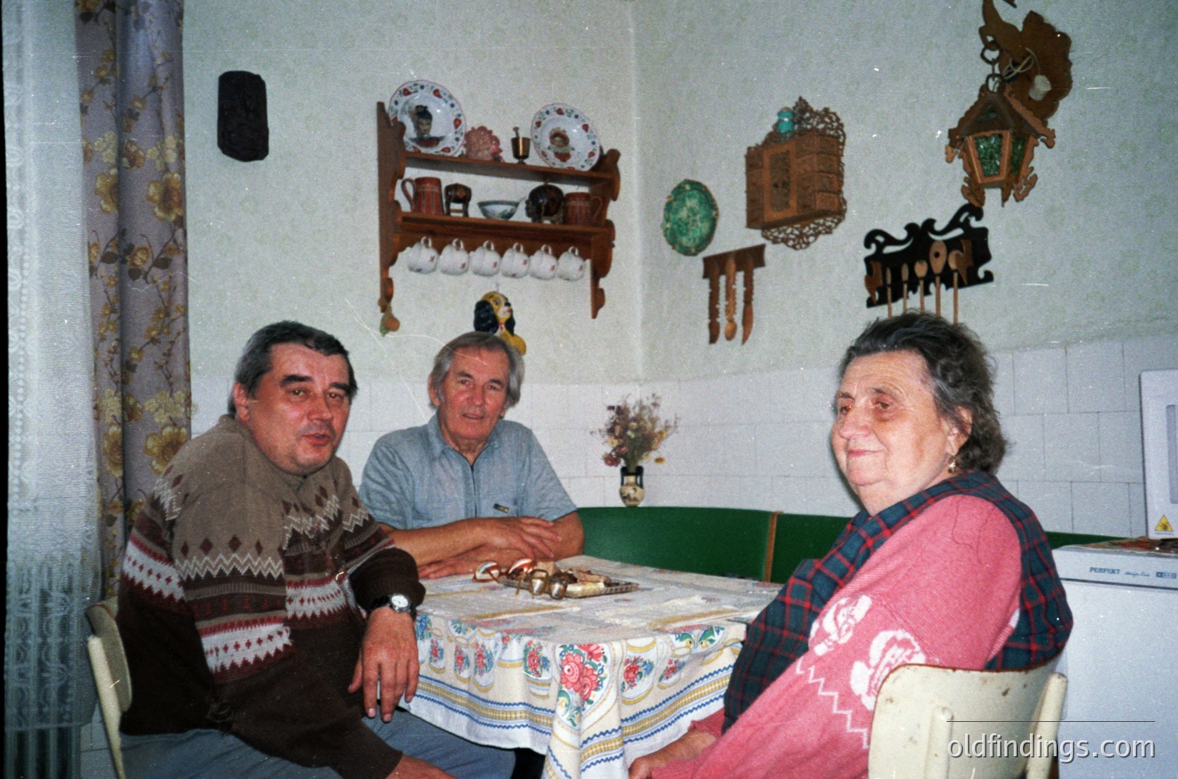 Three adults seated at a table in a modest indoor setting, likely a 1970s–1980s Eastern European home. The man on left wears a patterned sweater, center wears a light blue shirt, and woman on right wears a pink sweater with a checkered scarf. Tablecloth features floral patterns; shelves above display ceramic figurines and decorative items. Wall tiles and vintage decor suggest a lived-in, family-oriented space.