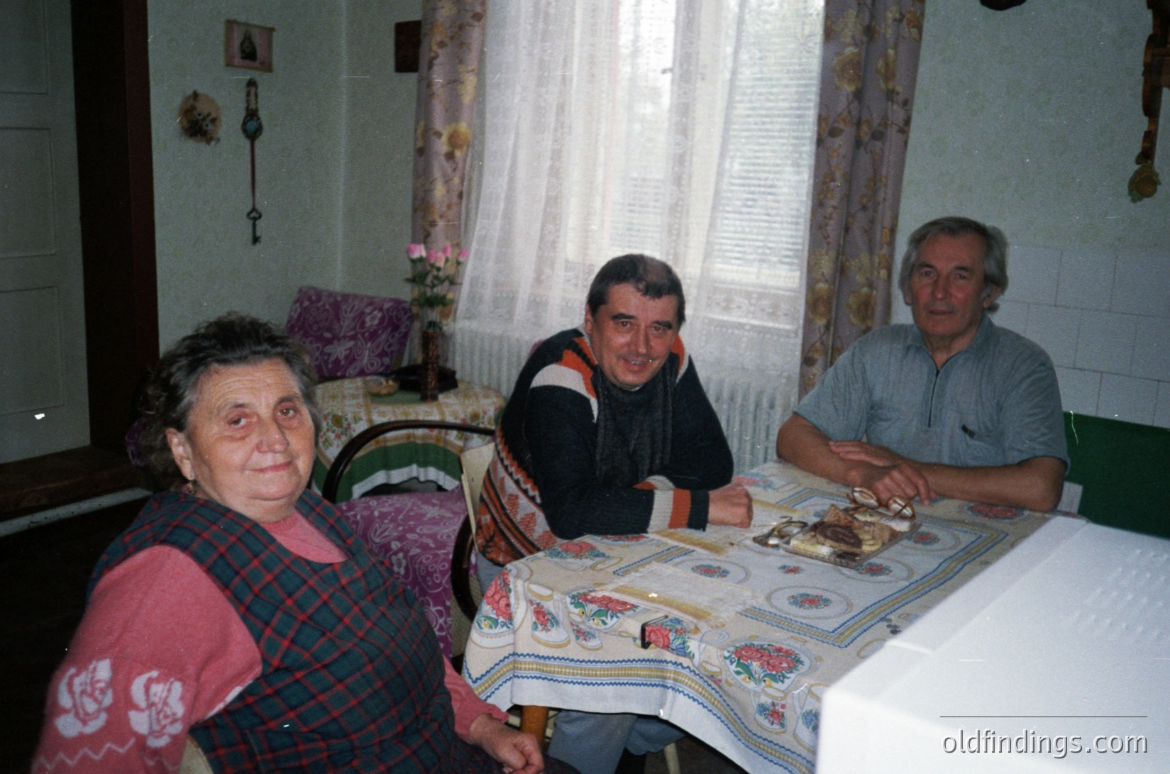 Three adults seated indoors at a table covered with a floral-patterned tablecloth, likely in a rural or traditional home setting. The woman wears a plaid sweater, the man in the center a striped sweater, and the man on the right a button-down shirt. A cake with candles sits on the table, suggesting a celebration. Decorative elements include a curtain with floral patterns, a wall-mounted candle holder, and a framed picture on the wall. The scene evokes a mid-20th century domestic atmosphere.
