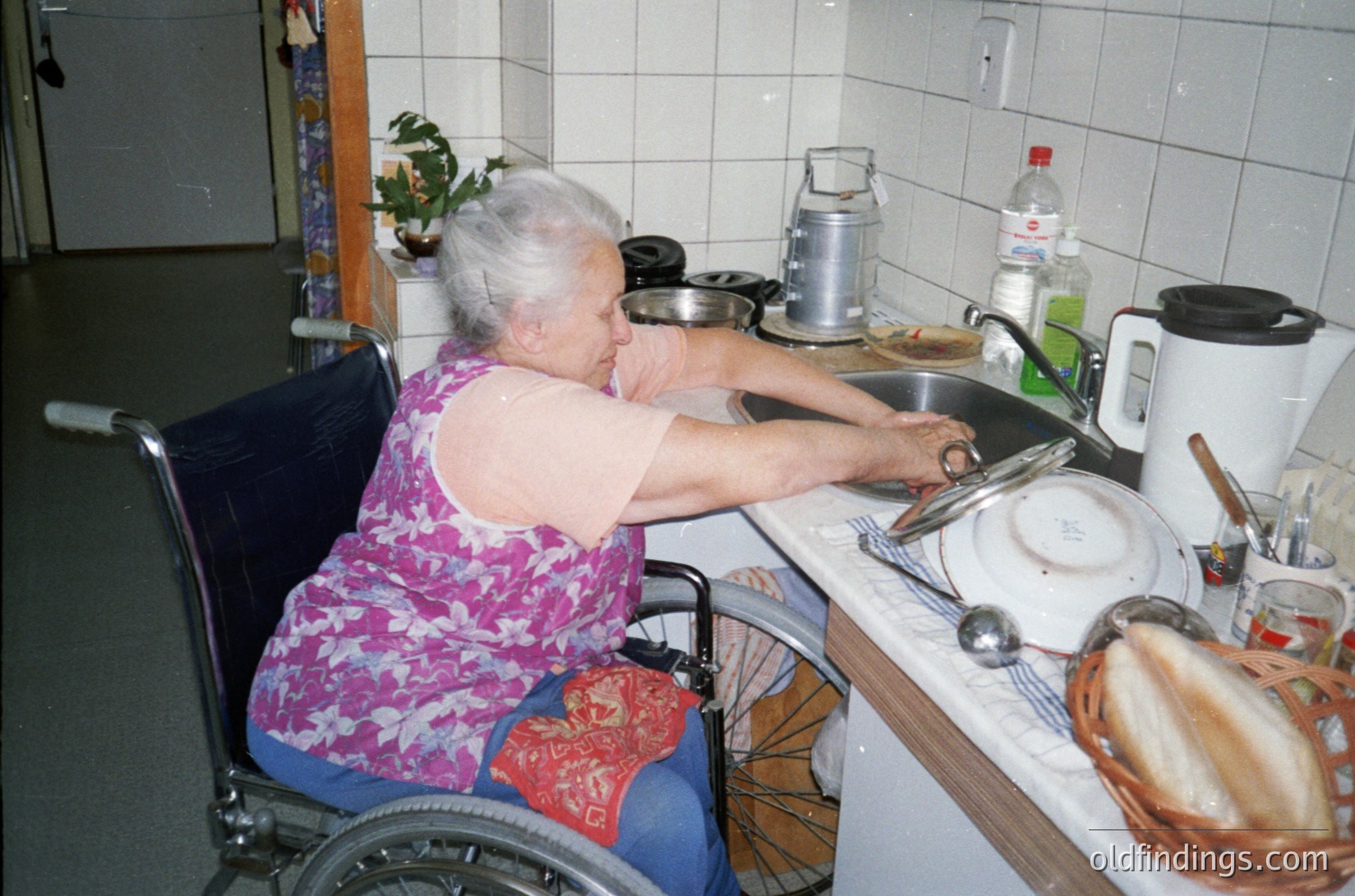 Indoor kitchen scene featuring an elderly woman in a wheelchair preparing food. She wears a floral-patterned sleeveless top and blue pants with red embroidered details. The kitchen includes white tiled walls, a sink with pots, a kettle, and a basket of bread. The setting suggests domestic life in a mid-20th-century home.