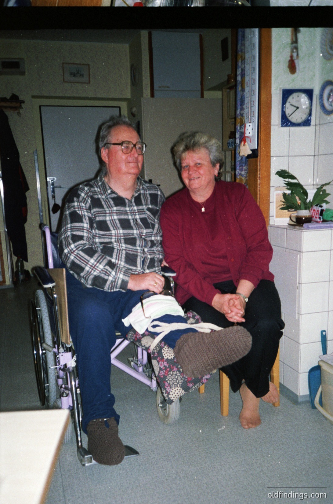 Two elderly individuals seated together in a clinical indoor setting, likely a healthcare facility. The man uses a wheelchair with a plaid shirt and glasses; the woman sits beside him in a maroon top. White tiled walls, a clock, and a potted plant in the background suggest a 1990s–2000s era. [Elderly couple in wheelchair, indoor healthcare setting, 1990s–2000s ]