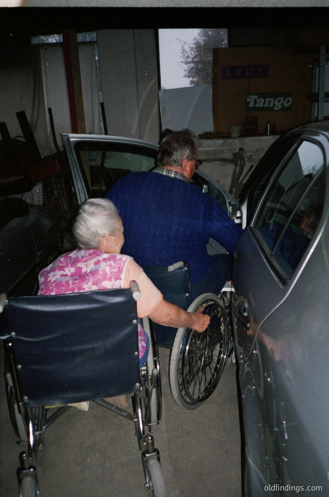 Couple assisting elderly woman into a car via wheelchair at a gas station with "Tango" branding. Indoor/outdoor lighting suggests evening. Mid-20th century automotive design and stationery signage indicate .