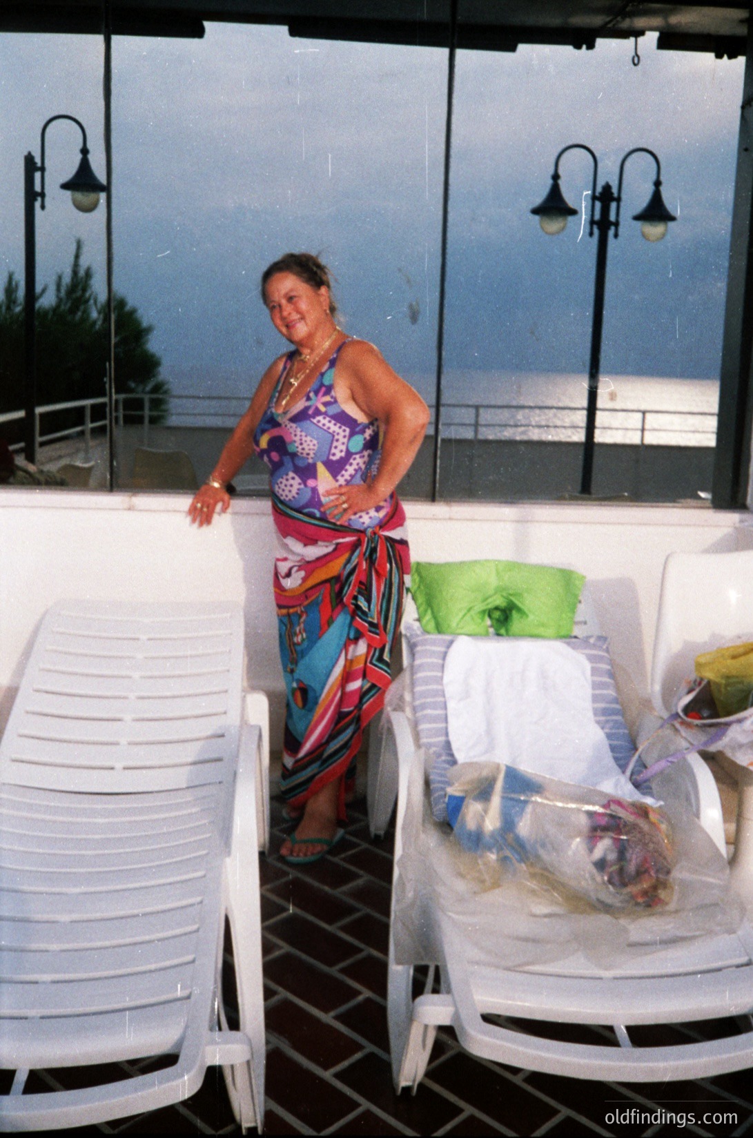 Brightly patterned sarong and tank top by seaside café patio, featuring vintage plastic chairs and lanterns. Overcast coastal setting suggests a Mediterranean or Black Sea locale, likely 1980s–1990s. Ideal for travel, hospitality, or vintage design references. é