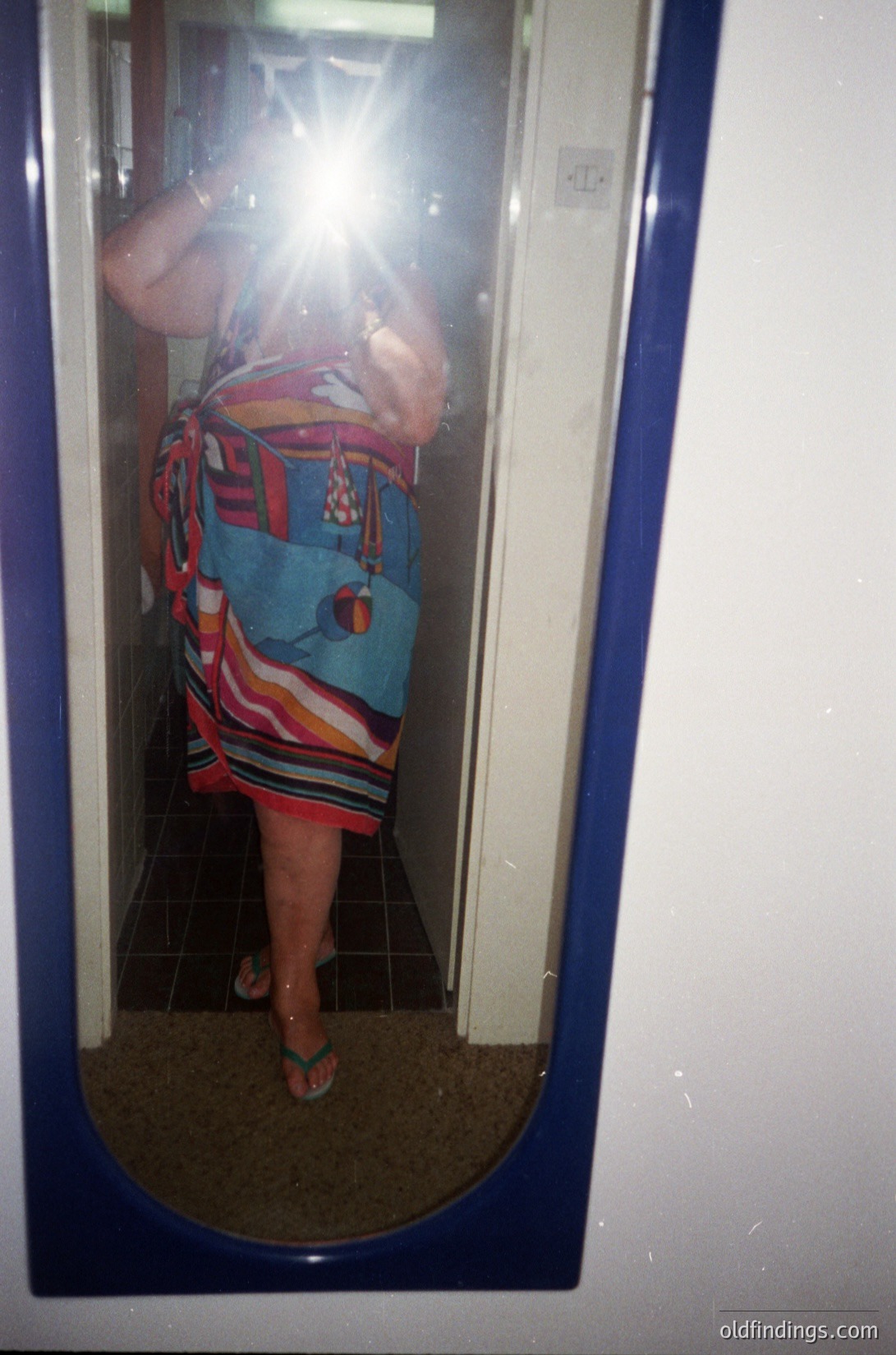 Vintage mirror selfie of a person in a bold, patterned dress with geometric and floral motifs in blues, reds, and whites. Reflective surface captures a doorway with blue trim and a light switch. Style suggests 1970s-1980s retro fashion.