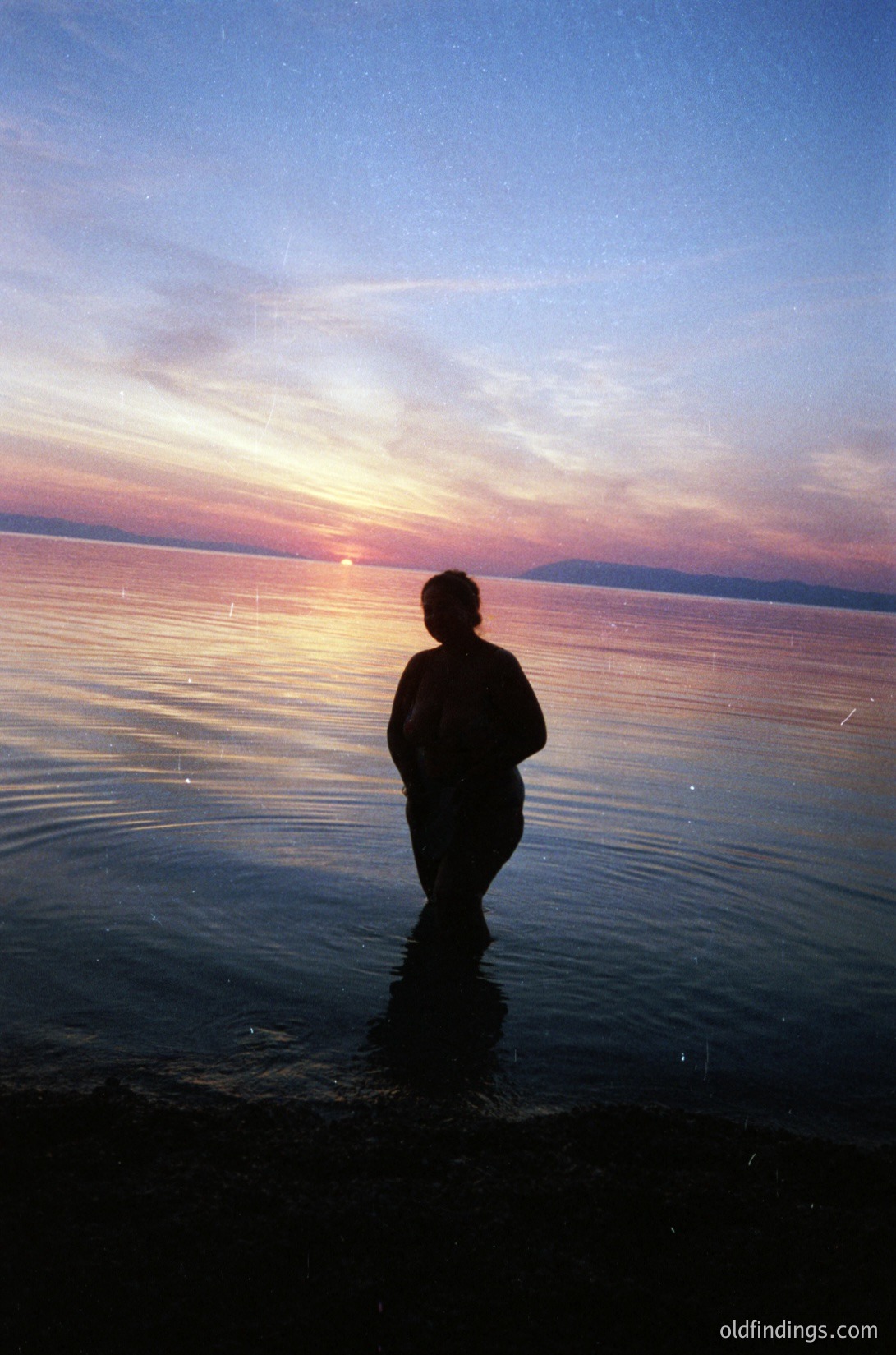 Silhouetted figure stands at water’s edge during golden-hour sunset, back to camera. Calm waters reflect fading sunlight; distant shoreline and hills faintly visible. Silhouette suggests mid-20th century clothing style.