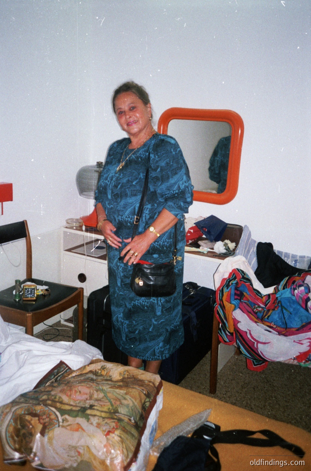 A woman in a patterned teal dress poses in a modest hotel room, likely from the **1990s**. She stands beside a wooden desk with a mirror, suitcase, and scattered clothing. The room’s utilitarian decor includes a bed with a floral-patterned coverlet and a small table lamp. The setting reflects mid-range hospitality of the era. [Mid-1990s hotel room with vintage decor, floral-patterned bedding, and patterned dress ]