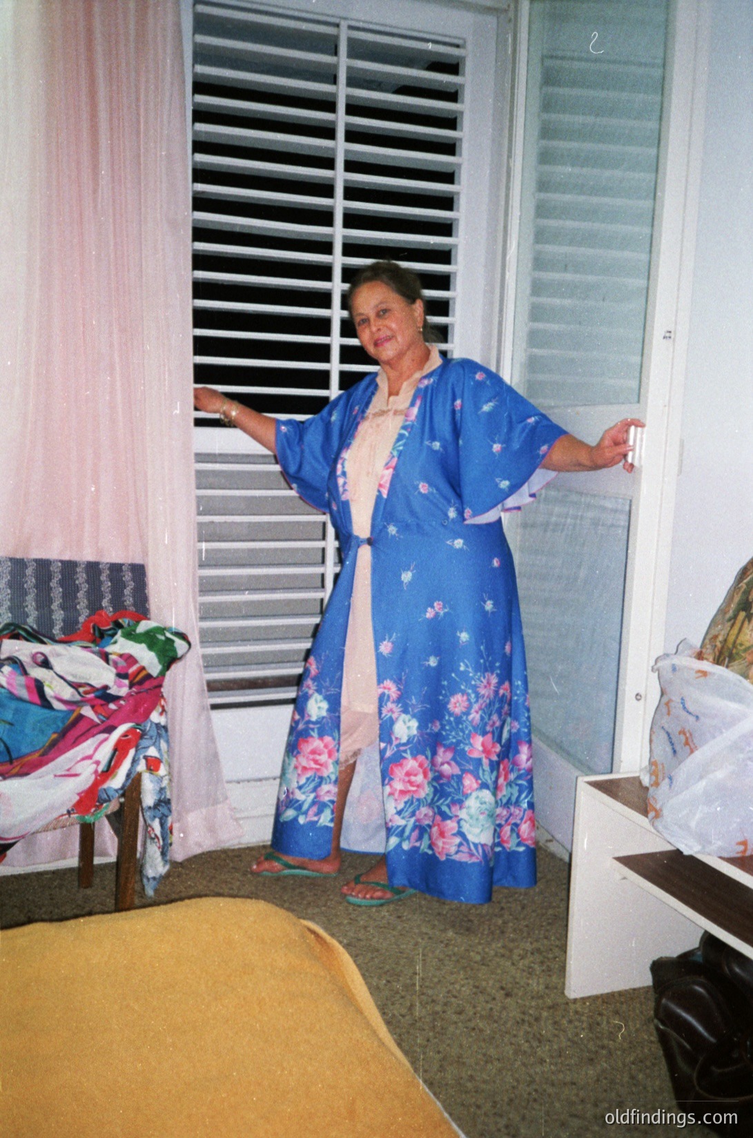 A woman in a floral-patterned blue robe poses indoors, leaning on a window frame with closed louvered blinds. She wears pink sandals and a light-colored underlayer. The room features patterned curtains, a striped pillow, and a partially visible sofa. Likely mid-20th century domestic setting.