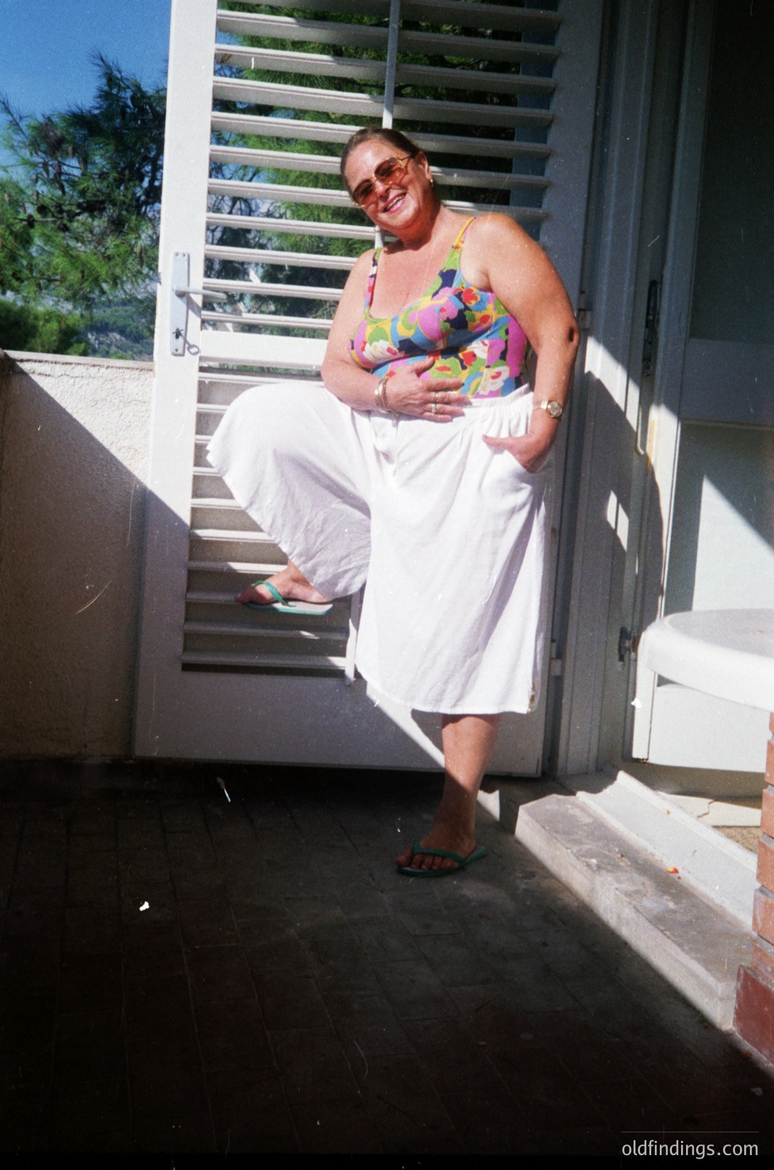 Vintage 1990s beachside portrait of a woman in a floral tank top and white pants, posing with a baby on a sunlit balcony. White railings and sliding door suggest a resort-style setting. Soft focus and warm lighting evoke nostalgia.